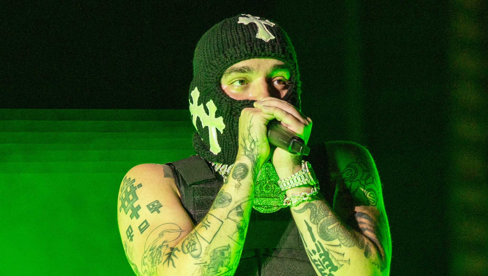 A person wearing a black ski mask with white cross designs, performing on stage with tattoos on their arms, holding a microphone.