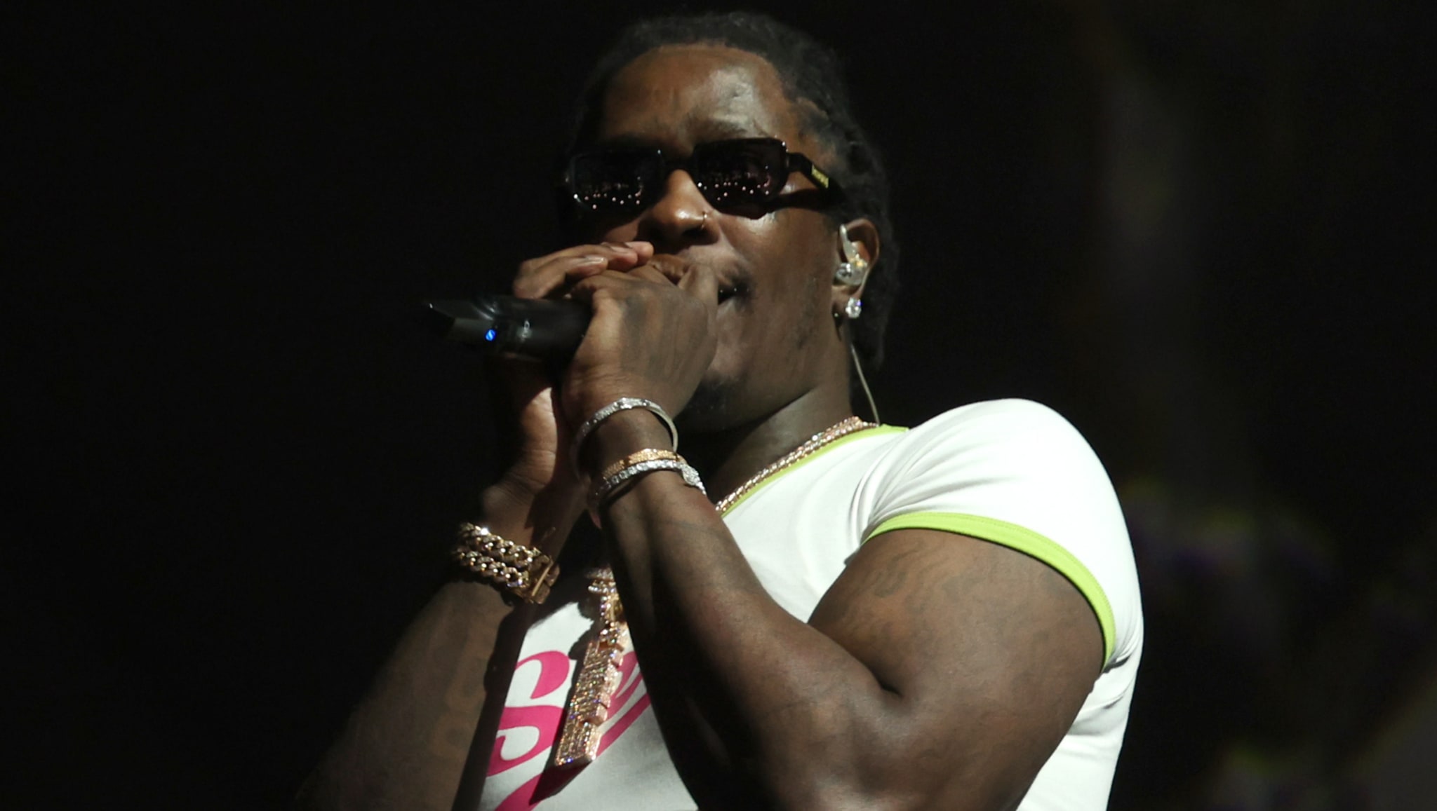 A performer wearing sunglasses and a white shirt sings into a microphone on stage.