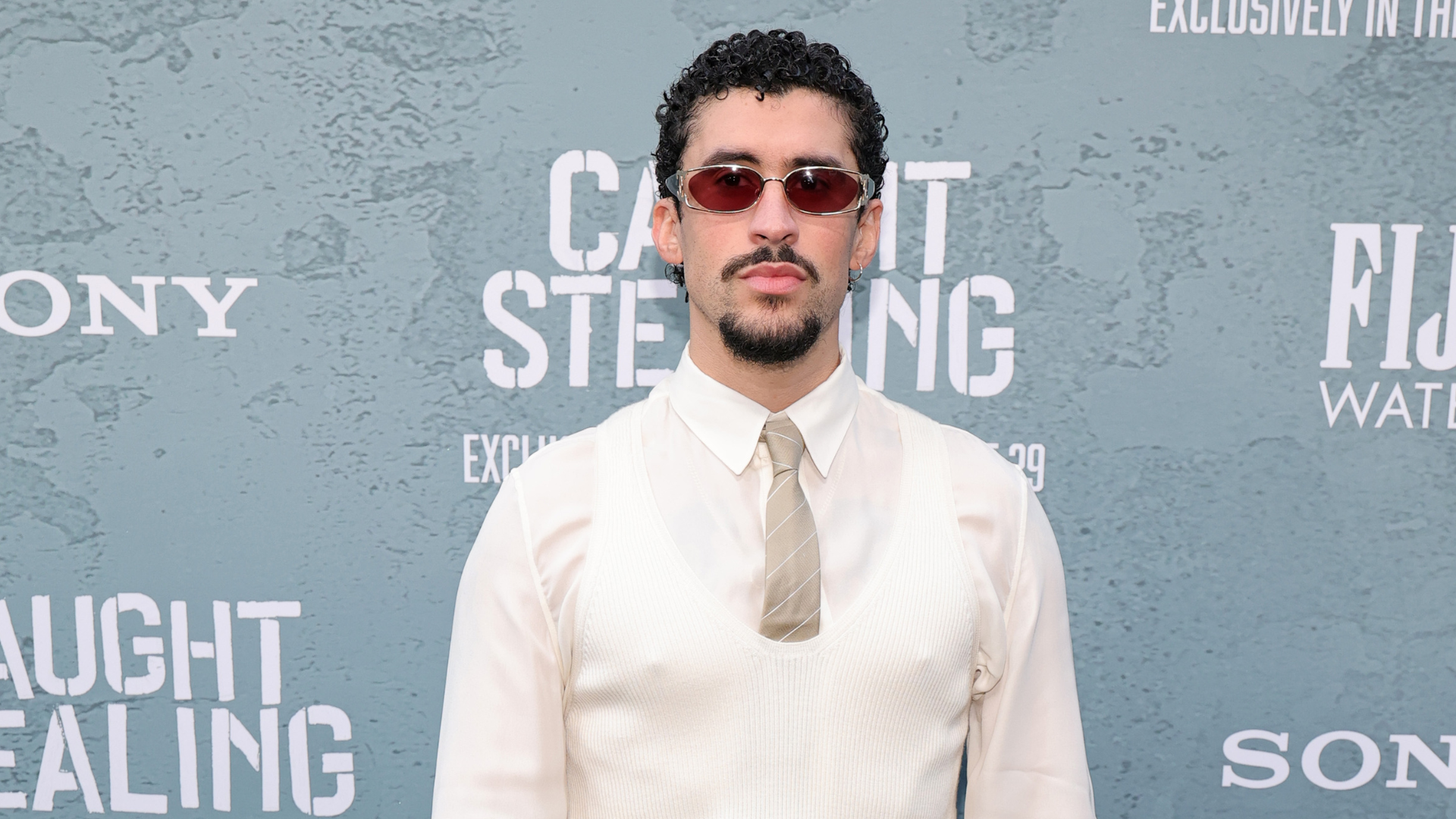 A man with curly hair and sunglasses stands in front of a backdrop with text, wearing a white shirt, tie, and vest.