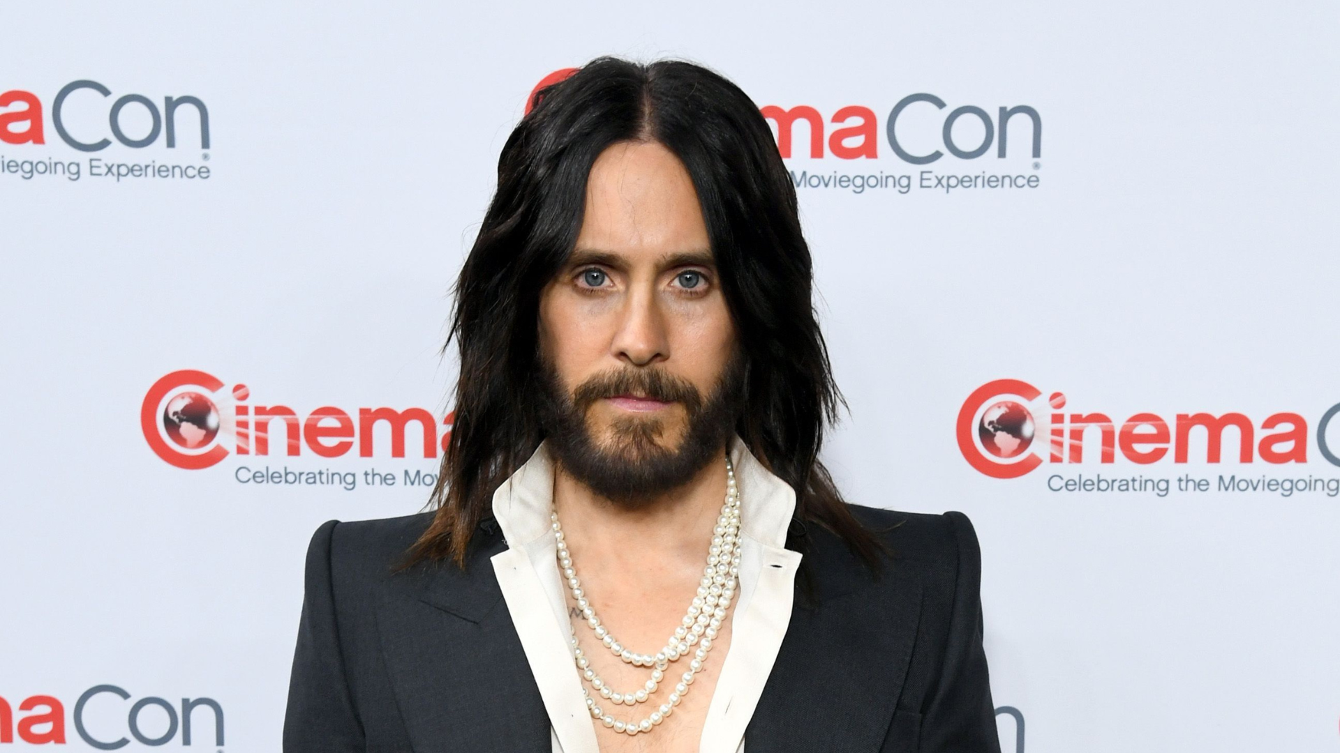 Jared Leto at CinemaCon, wearing a dark suit with a white shirt and layered pearl necklaces.