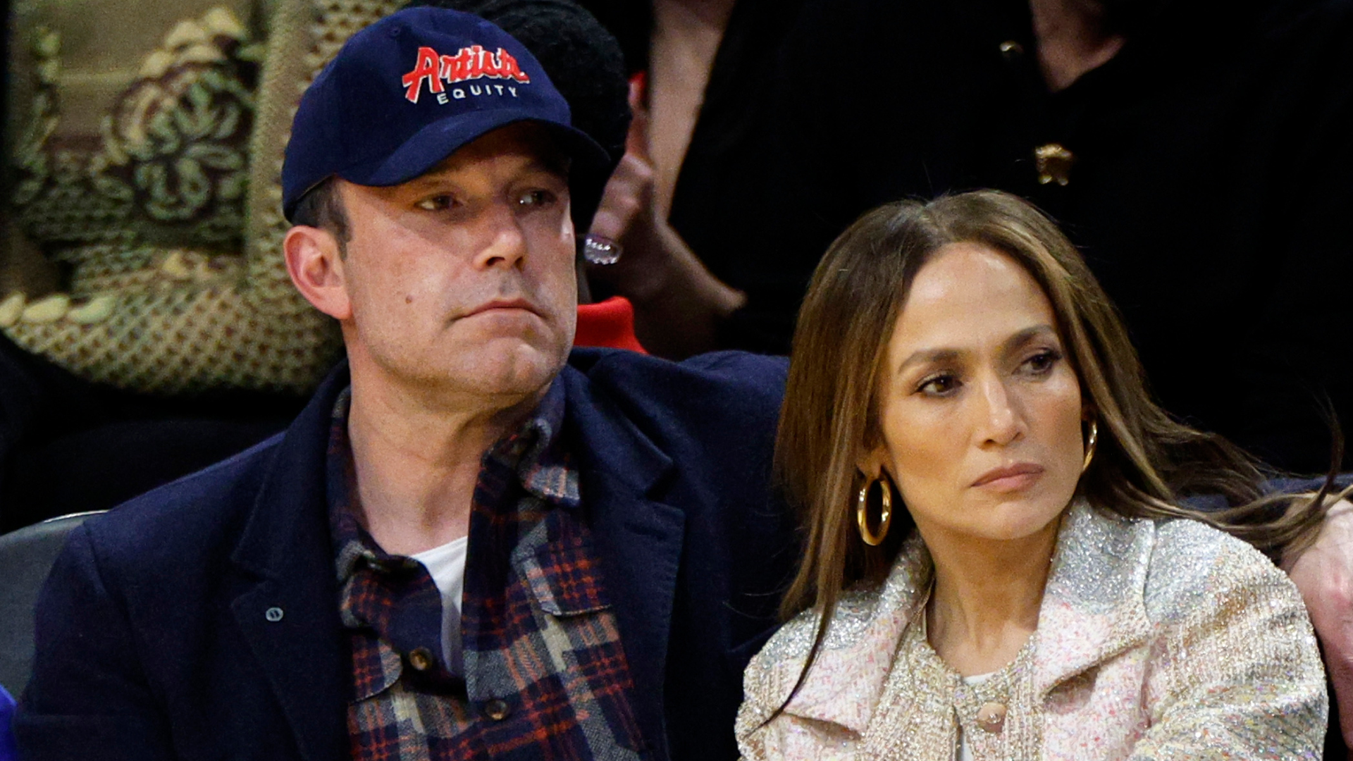 Ben Affleck and Jennifer Lopez sitting together at an event, with Affleck in a cap and Lopez in a light-colored outfit.