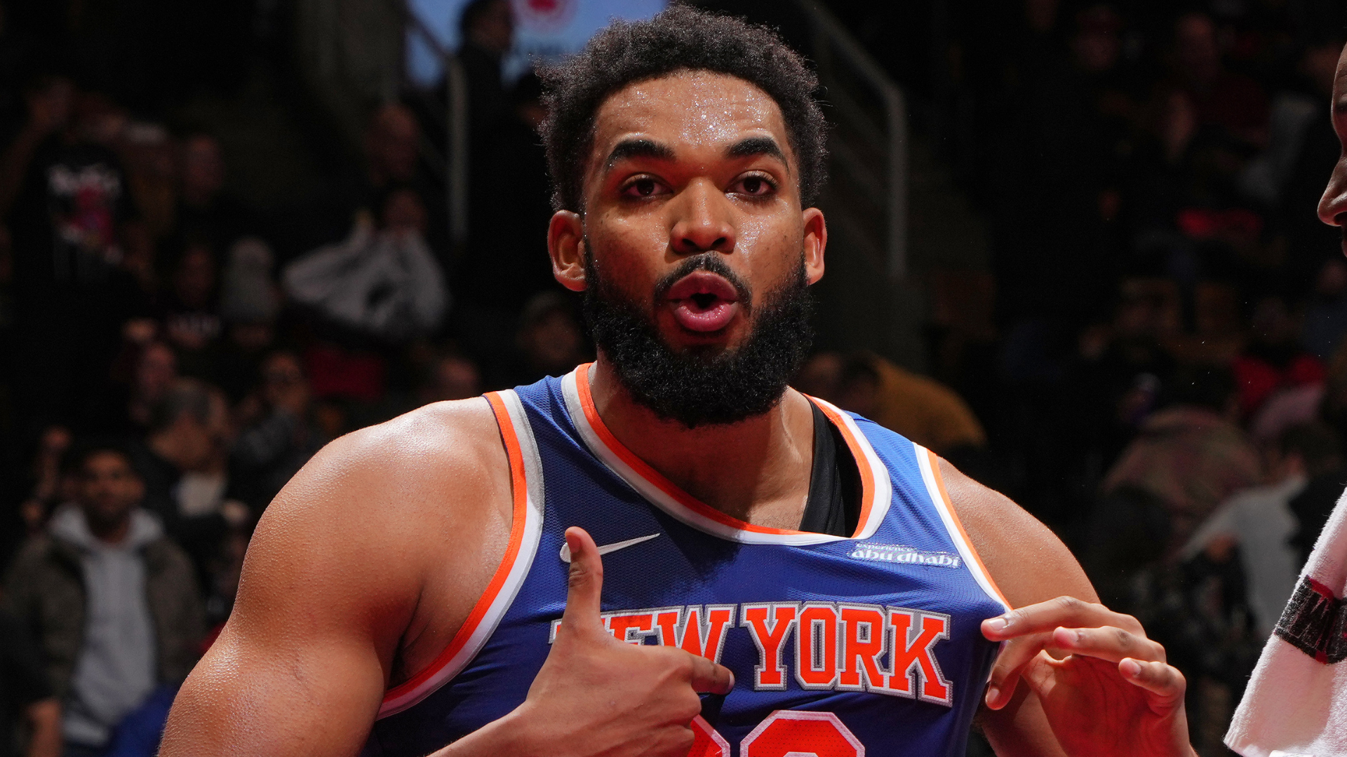 Basketball player in a New York jersey on the court, gesturing with his hands, with a crowd in the background.
