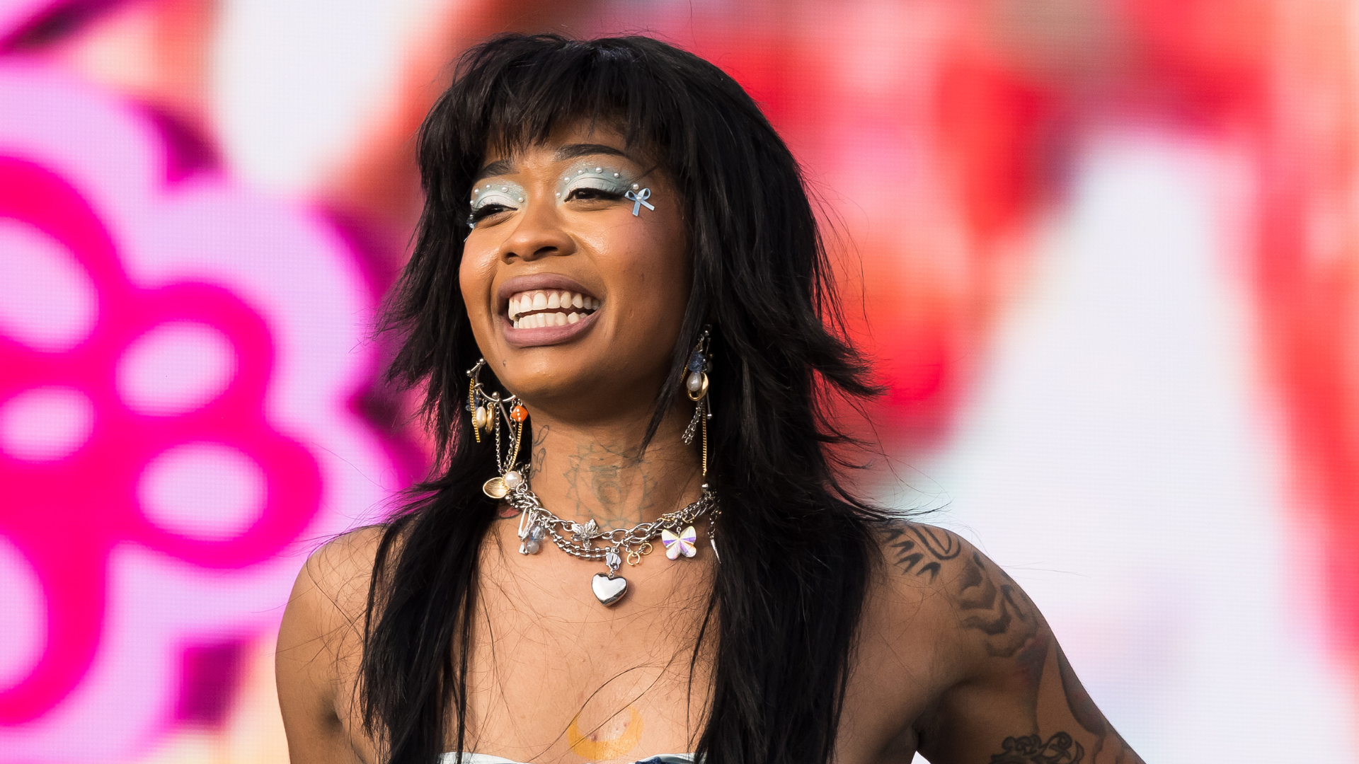 A person with long dark hair, wearing silver eye makeup and a necklace with charms, smiles against a colorful background.