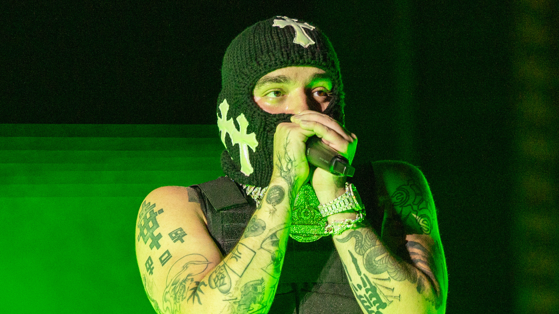 A person wearing a black ski mask with white cross designs, performing on stage with tattoos on their arms, holding a microphone.