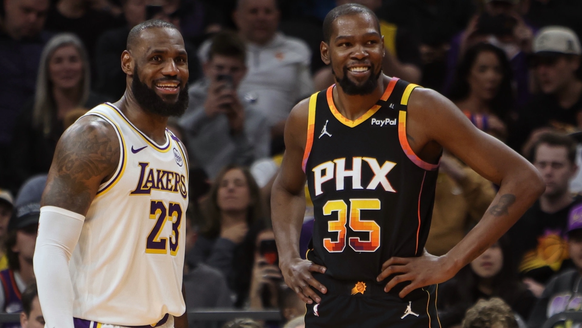 LeBron James in a Lakers jersey and Kevin Durant in a Suns jersey smiling on a basketball court.