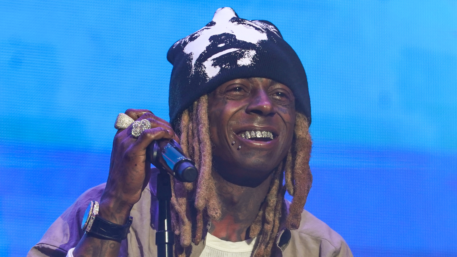 Lil Wayne performing on stage, wearing a black beanie, with dreadlocks and diamond-studded grillz, holding a microphone.