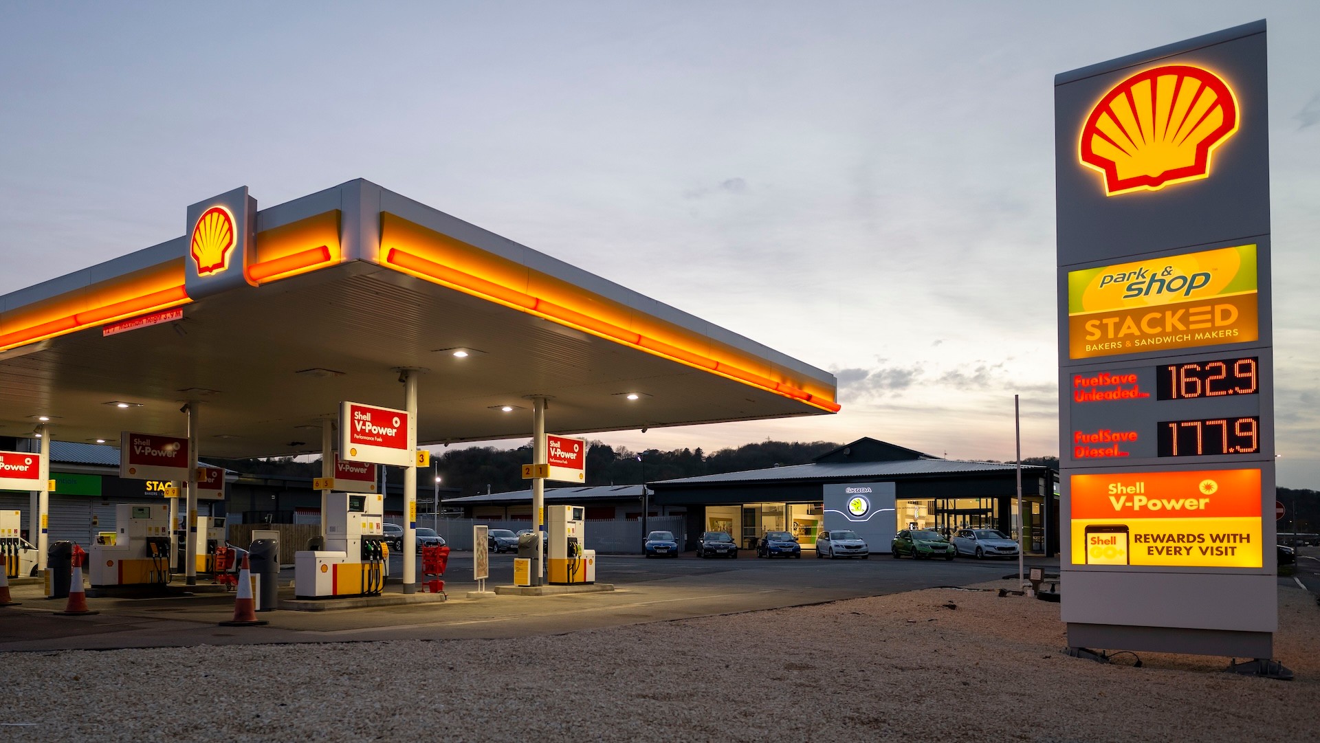 A Shell gas station at dusk, with illuminated signage displaying fuel prices. The forecourt is empty, and a convenience store is visible.
