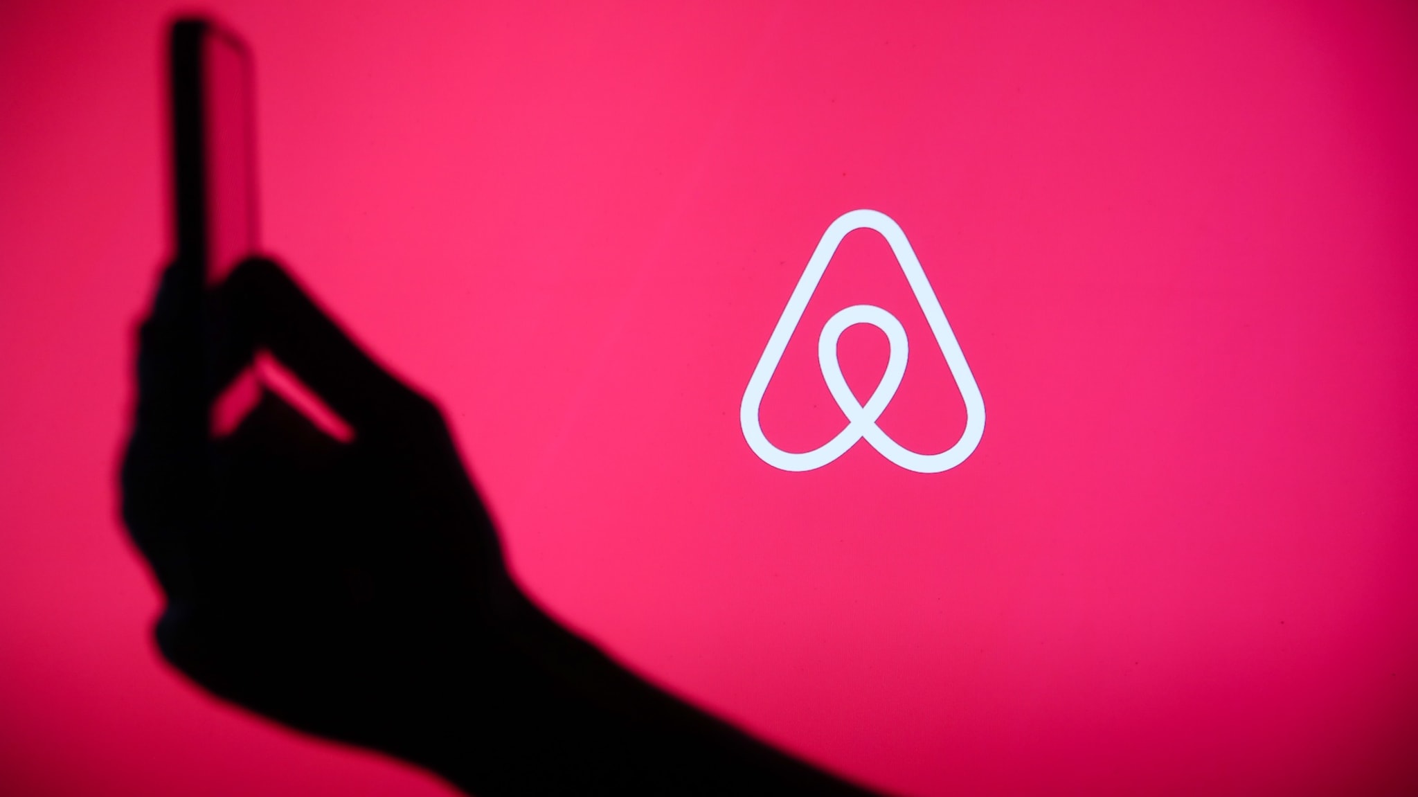 Silhouette of a hand holding a phone against a pink background with the Airbnb logo.