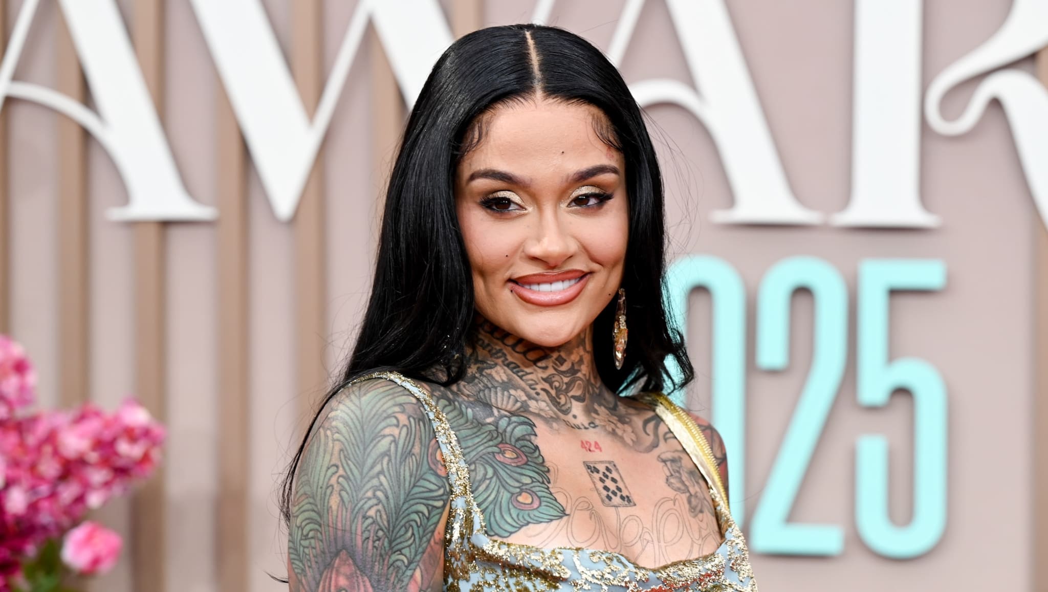 A woman with long black hair and extensive tattoos, wearing a gold dress, smiles at an event.