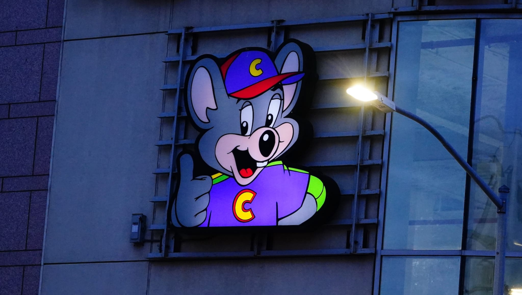 A sign featuring Chuck E. Cheese, a cartoon mouse character, wearing a cap and shirt with the letter "C," giving a thumbs up.