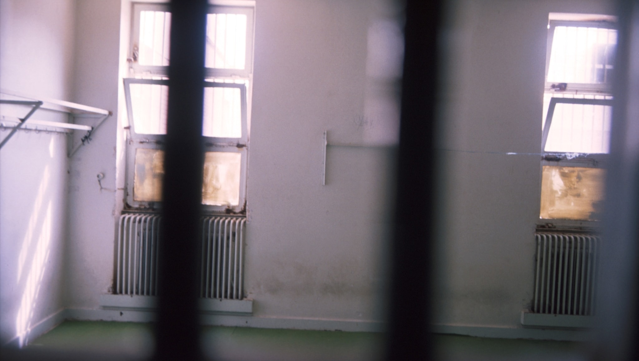 A dimly lit, empty room with barred windows and radiators beneath them. The view is partially obscured by vertical bars in the foreground.