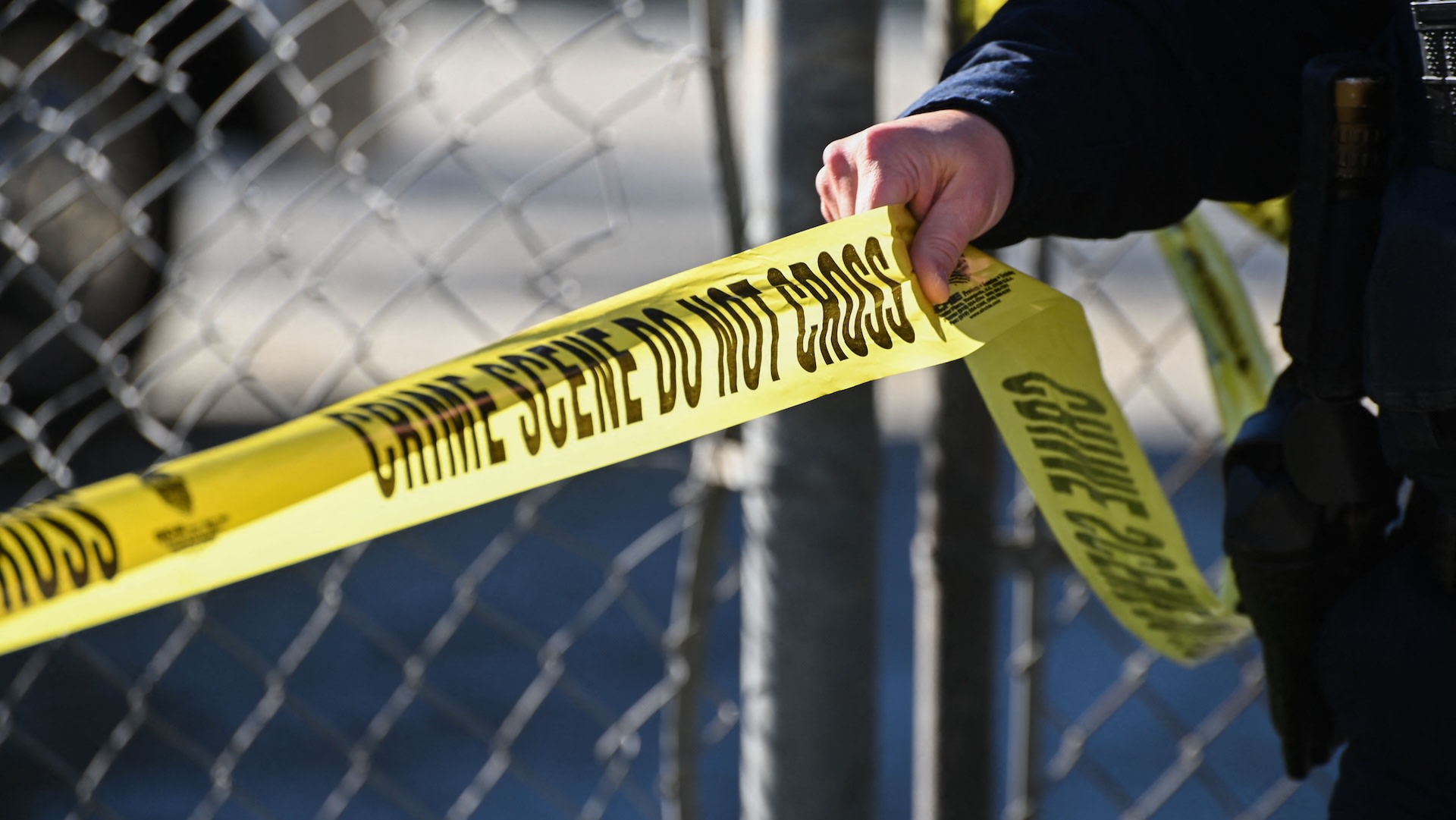 A police officer holding yellow "Crime Scene Do Not Cross" tape near a chain-link fence.