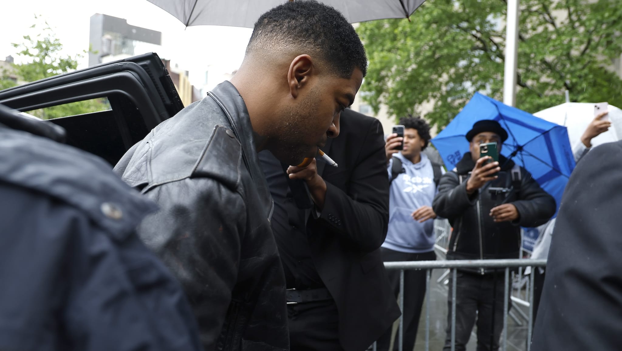 Kid Cudi in a leather jacket is surrounded by people with umbrellas. Others are taking photos with their phones.