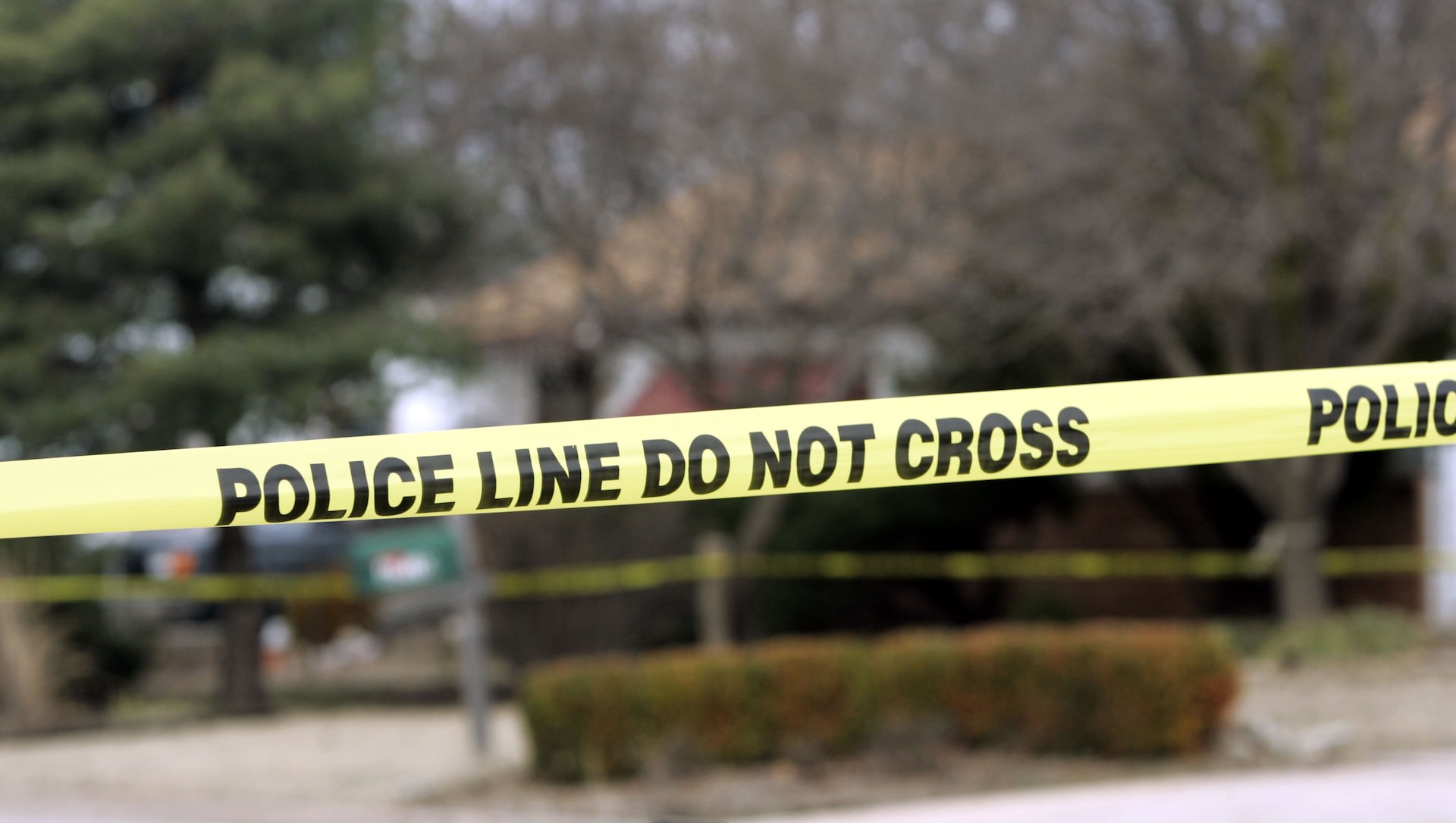 Yellow police tape with "POLICE LINE DO NOT CROSS" text, in front of a blurred outdoor scene with trees and a building.