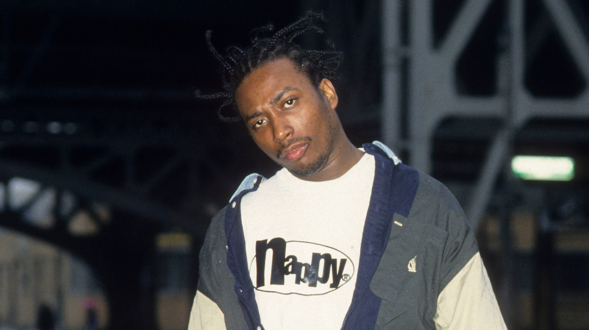 Rapper Ol' Dirty Bastard stands on a street wearing a layered shirt, baggy denim shorts, and sneakers, with a casual pose.