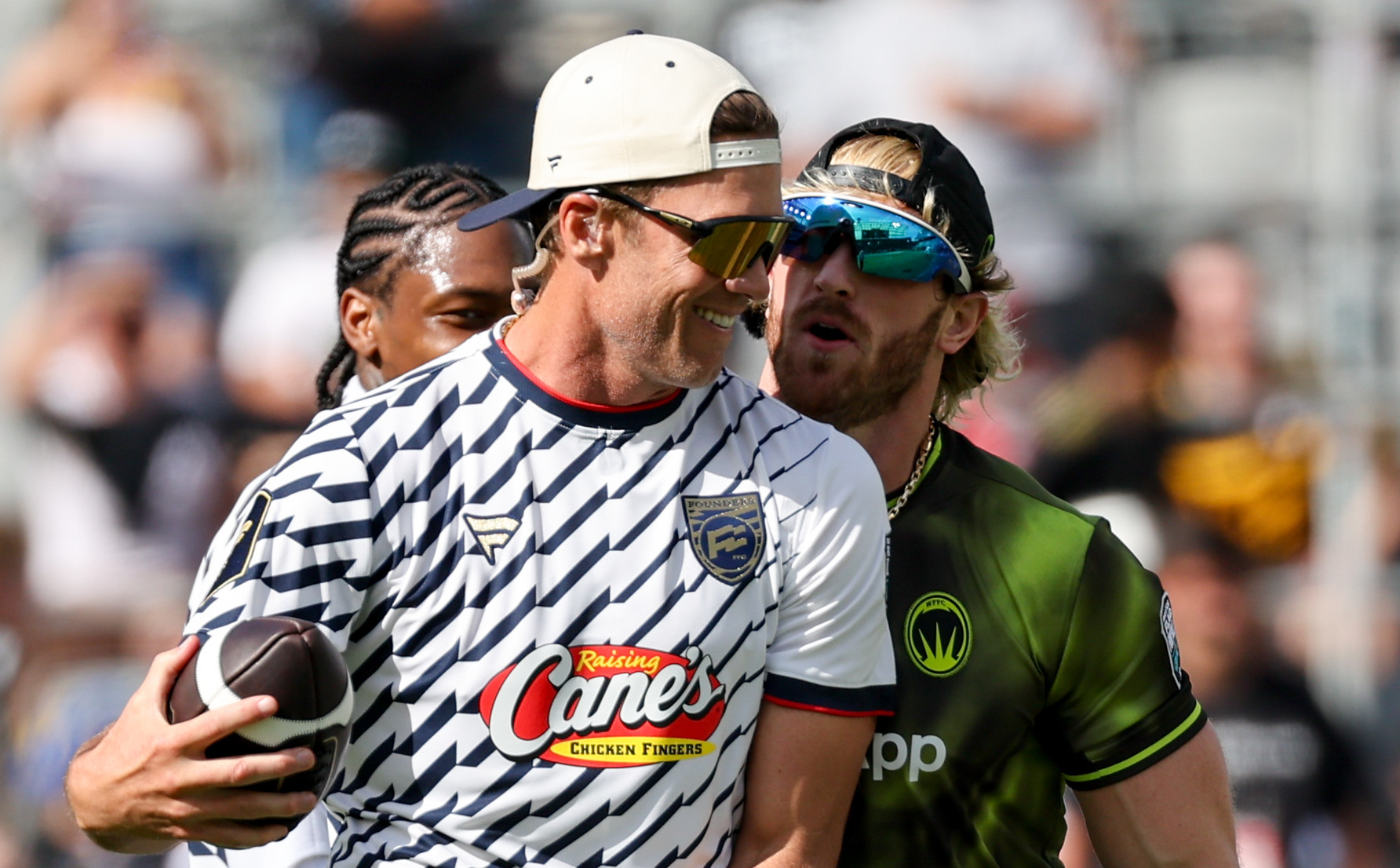 Tom Brady holding a football and wearing a cap, Logan Paul in sunglasses and a green jersey.
