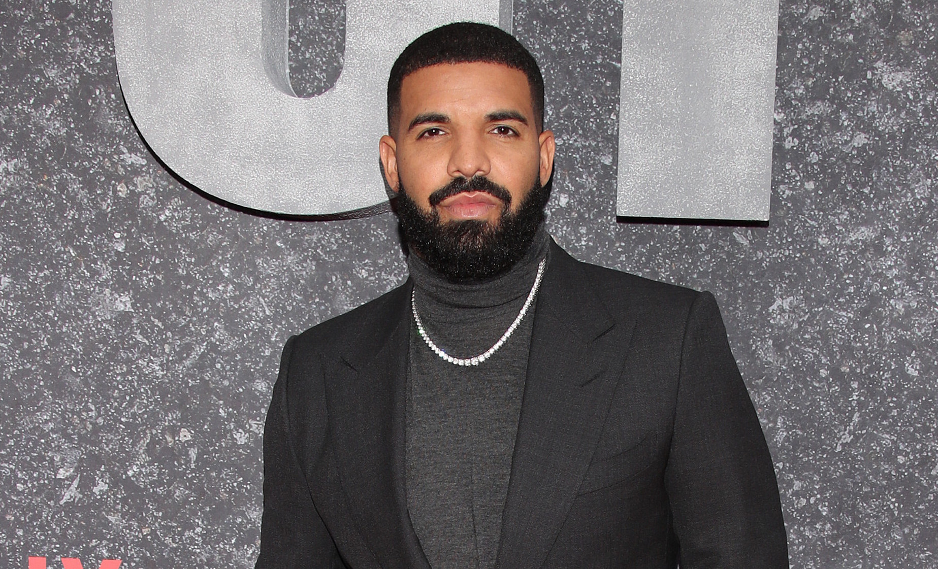 Drake at the "Top Boy" premiere, wearing a dark suit and turtleneck, stands on a red carpet with a Netflix backdrop.