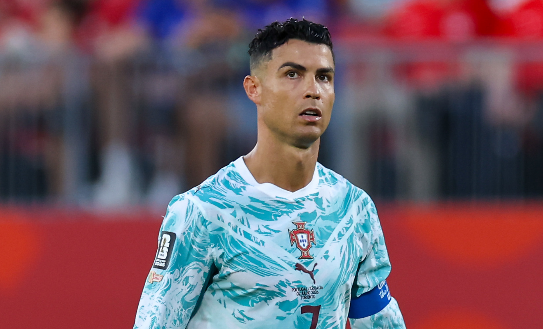 Cristiano Ronaldo in a Portugal jersey on the soccer field, wearing a captain's armband, looking focused.