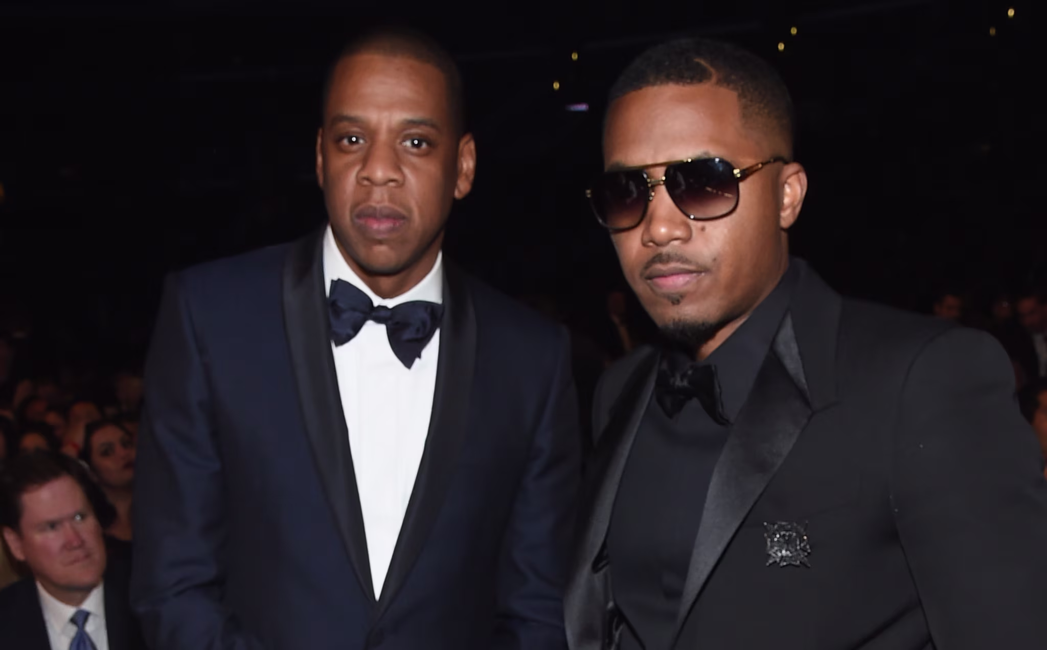 Jay-Z and Nas dressed in formal suits, standing together at an event. Jay-Z wears a blue tuxedo, and Nas wears sunglasses with a black suit.