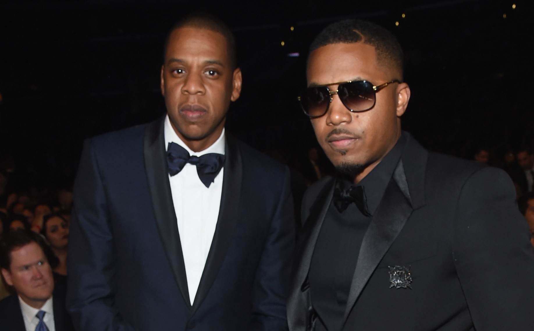 Jay-Z and Nas dressed in formal suits, standing together at an event. Jay-Z wears a blue tuxedo, and Nas wears sunglasses with a black suit.