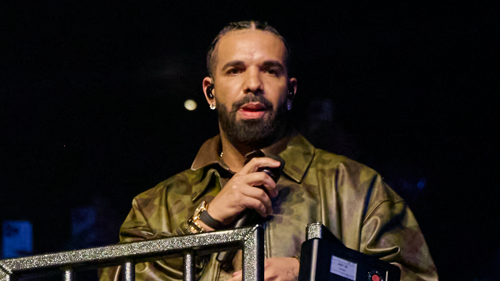Drake, wearing a camouflage jacket, holds a microphone on stage. He has braided hair and a beard.