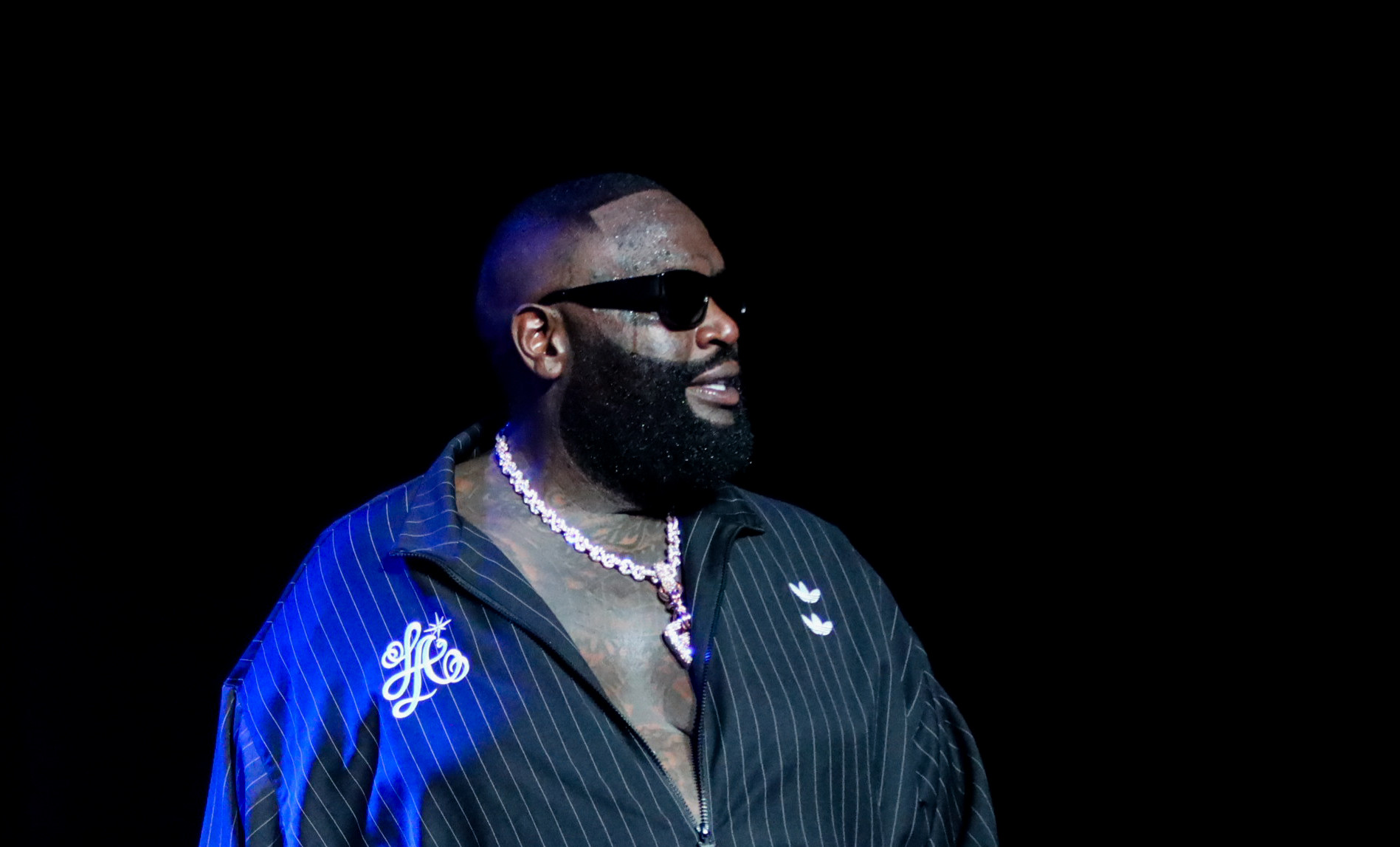 A man wearing sunglasses and a striped tracksuit stands on stage, illuminated by blue lighting.