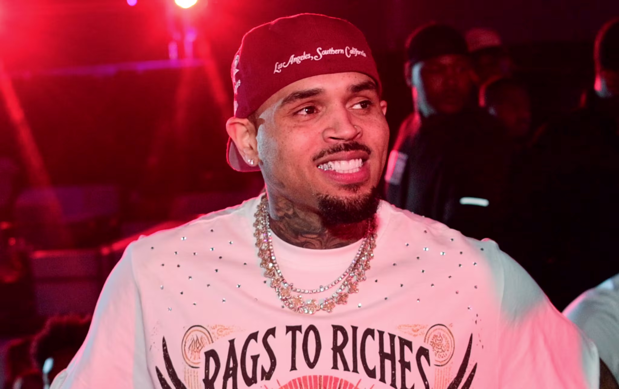 Chris Brown wearing a "Rags to Riches" shirt and a cap, standing in a dimly lit venue with red lighting.