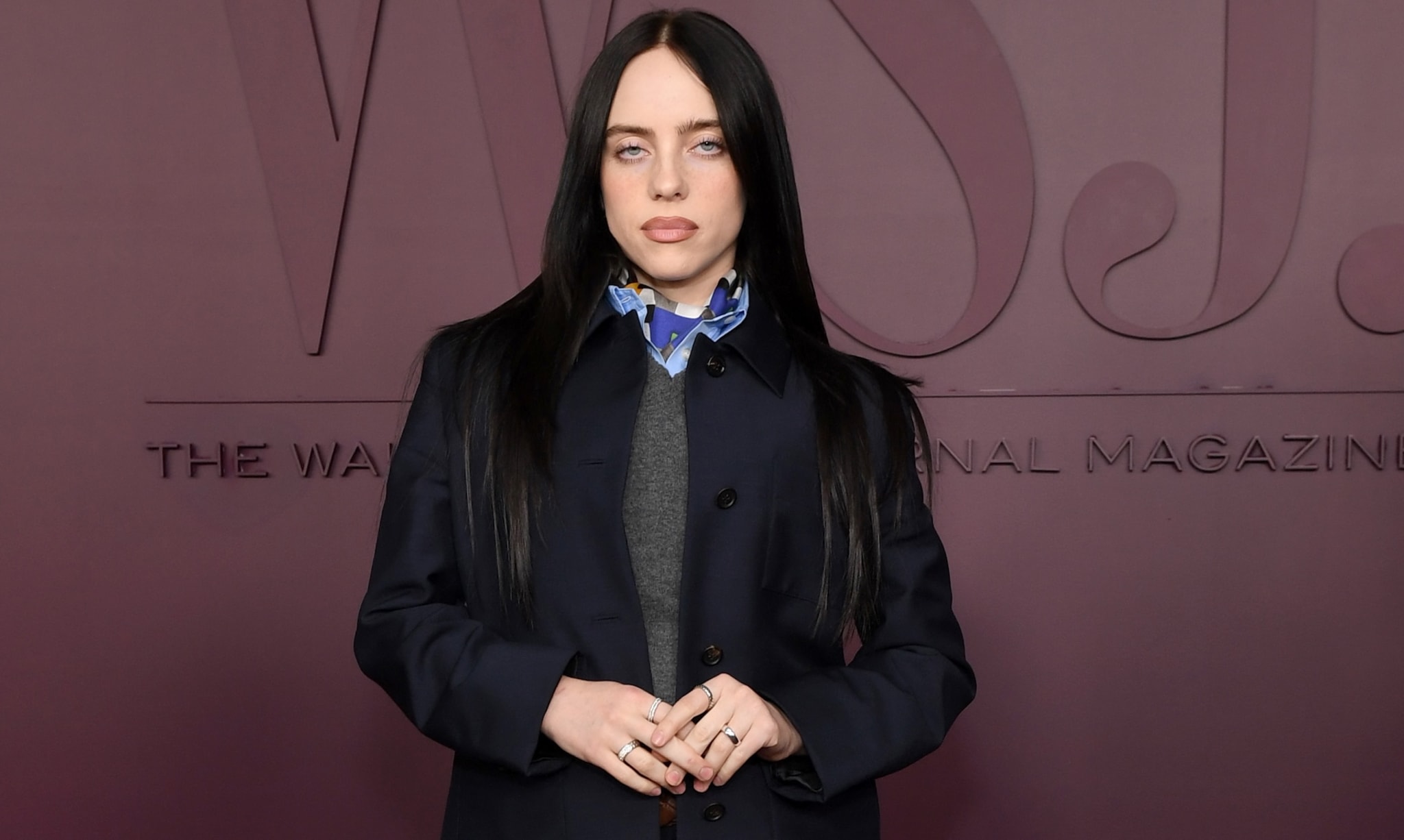 Billie Eilish at an event, wearing a dark coat over a gray sweater with a patterned collar, standing against a maroon backdrop.