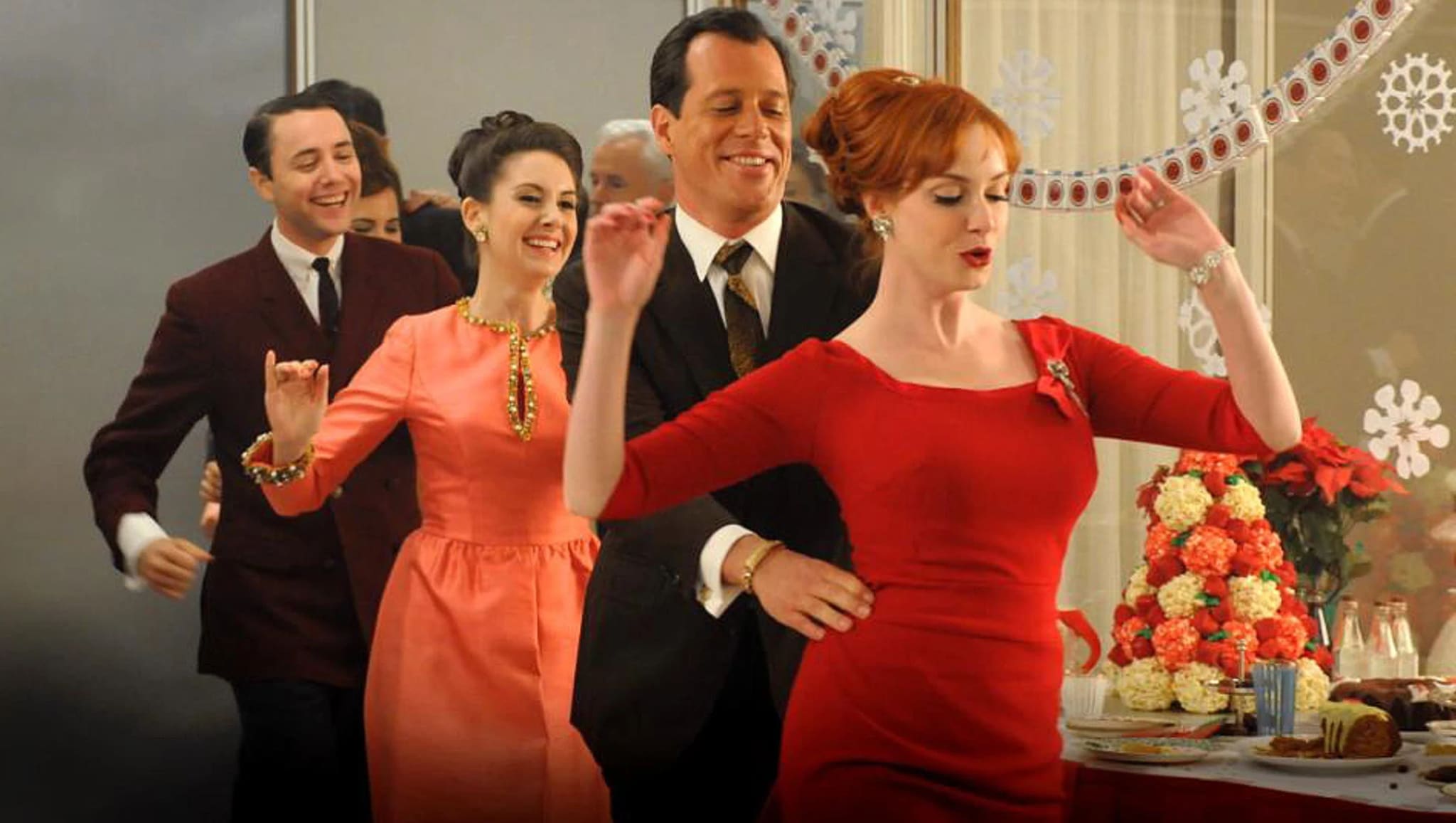 A festive office party scene with people dancing in a conga line. A woman in a red dress leads, with decorations and a table of food nearby.