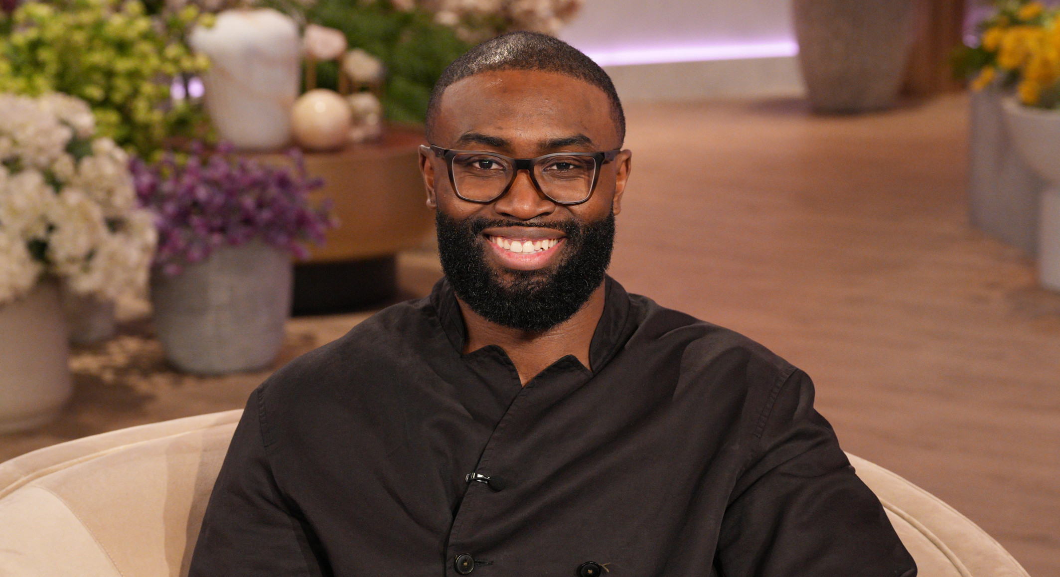 Jaylen Brown with glasses and a beard sits in a cozy chair, surrounded by colorful flowers in a warmly lit room.