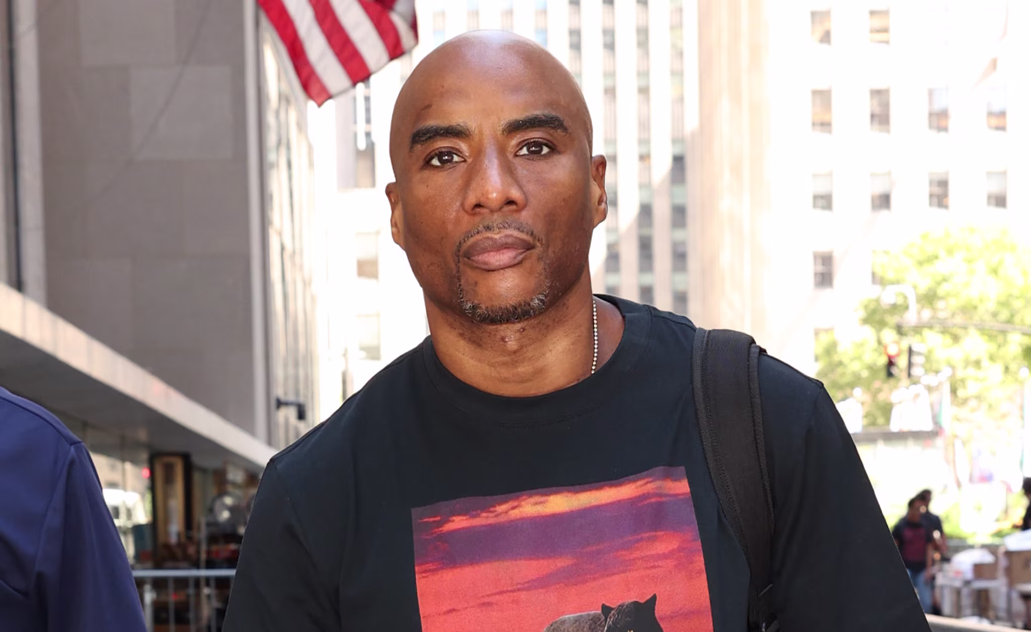 Charlamagne Tha God in a black T-shirt with a bear print, holding shoes, standing on a city street with American flag in the background.
