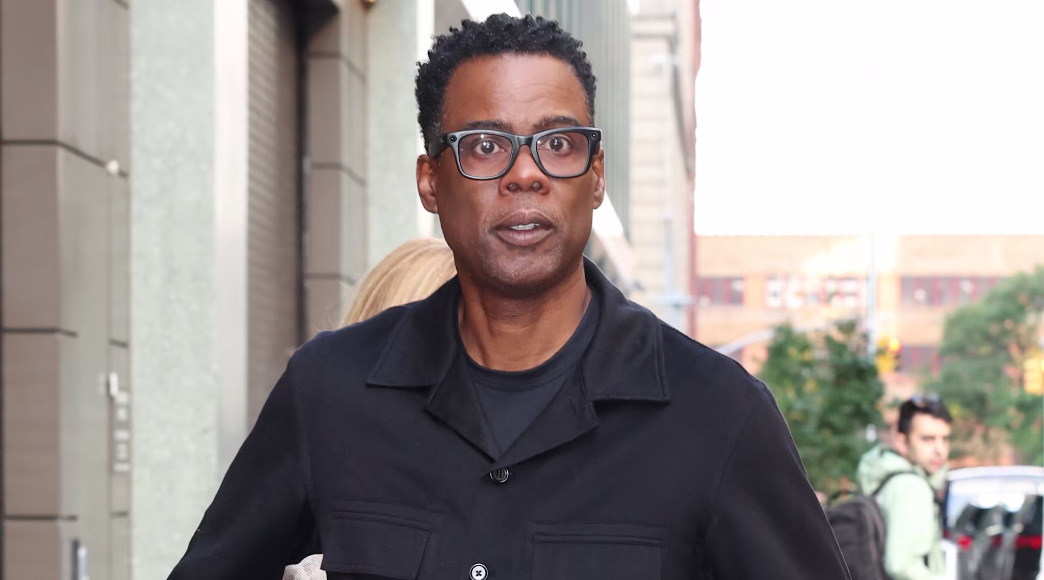 Chris Rock wearing glasses and a black outfit, walking on a city sidewalk.