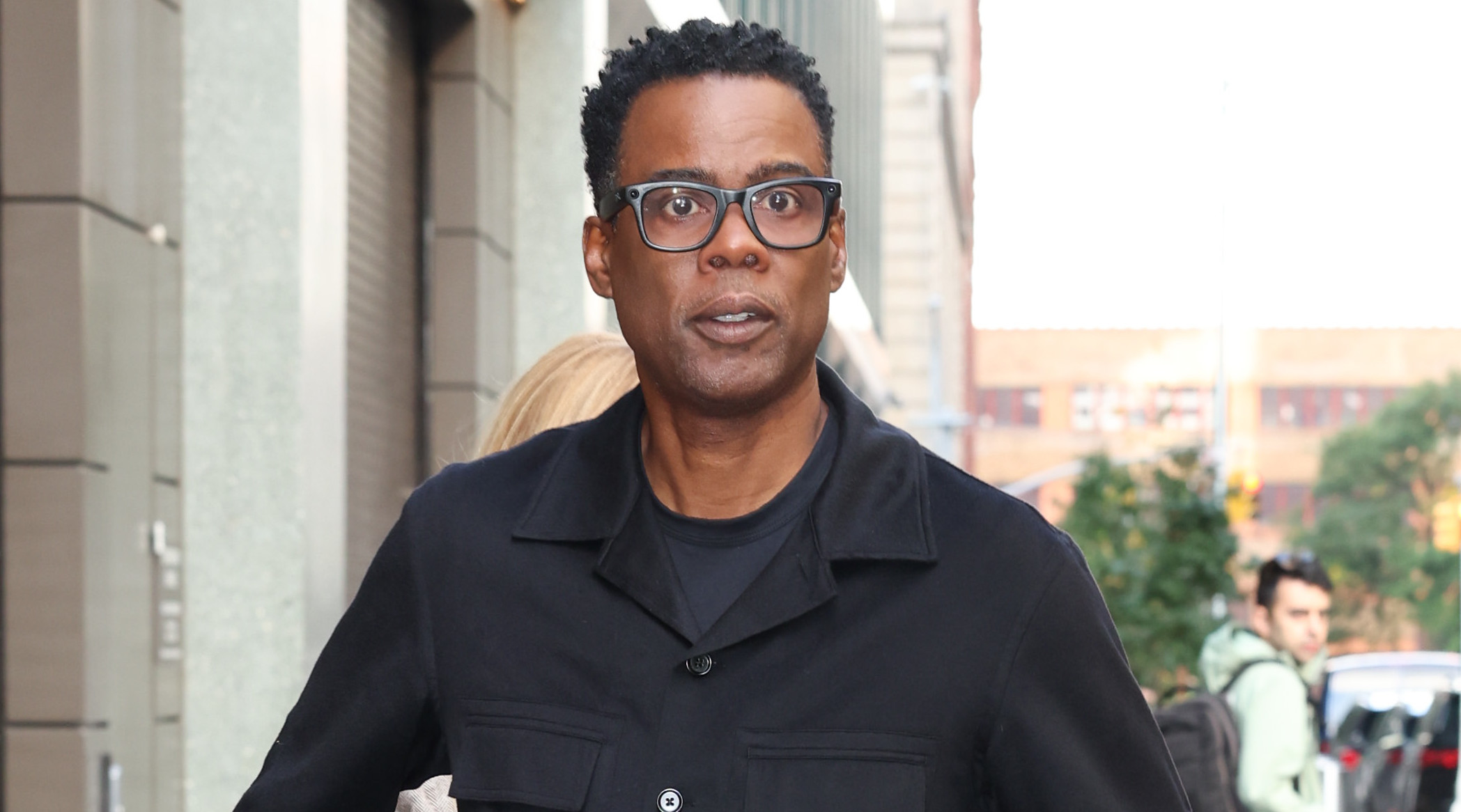 Chris Rock wearing glasses and a black outfit, walking on a city sidewalk.