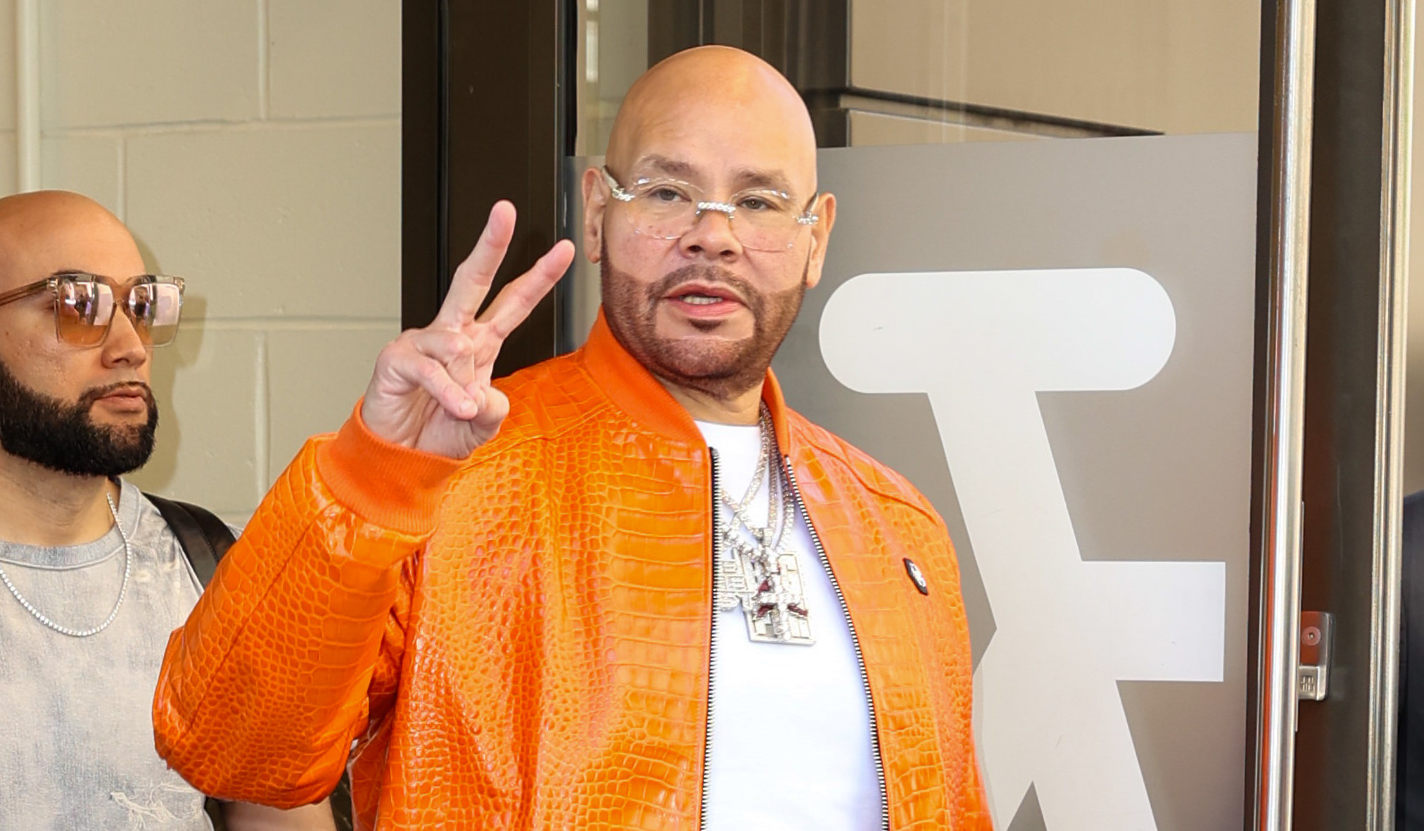 Fat Joe in an orange jacket making a peace sign while exiting a building, accompanied by two others.