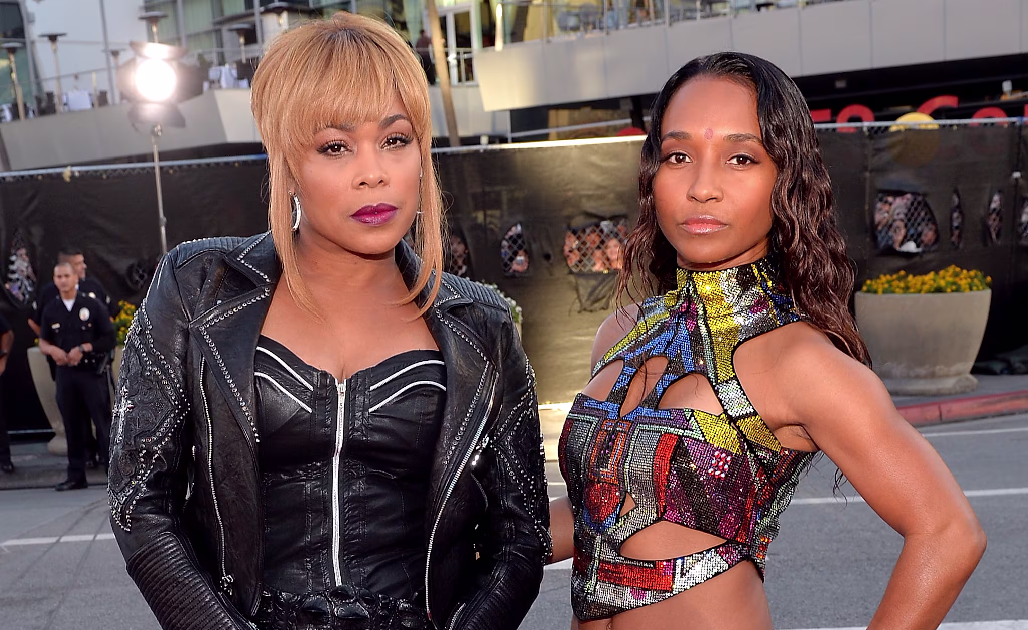 Tionne "T-Boz" Watkins and Rozonda "Chilli" Thomas from TLC pose on a street. T-Boz wears a black outfit; Chilli wears a colorful top and blue skirt.