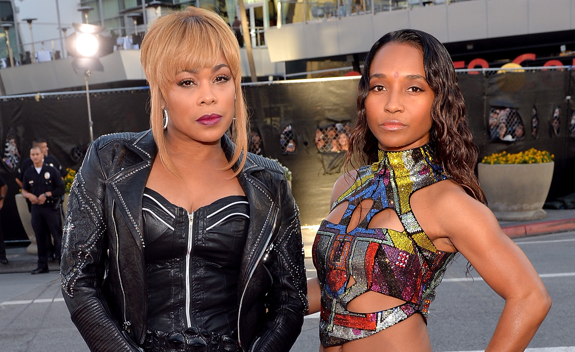 Tionne "T-Boz" Watkins and Rozonda "Chilli" Thomas of TLC pose on a street, with T-Boz in a black outfit and Chilli in a colorful top and blue skirt.