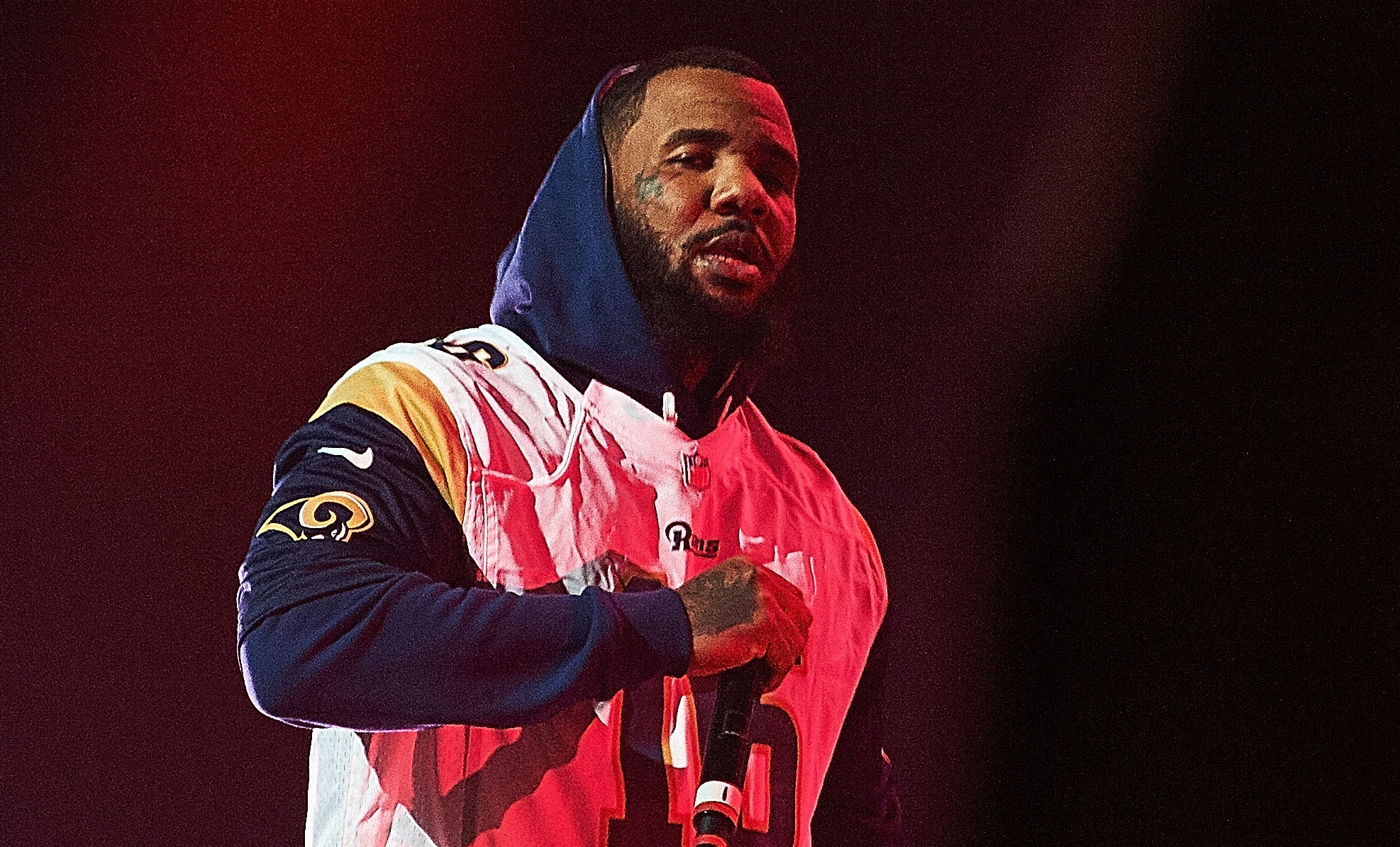 The Game performing on stage wearing a Los Angeles Rams jersey with a hood, under red stage lighting.