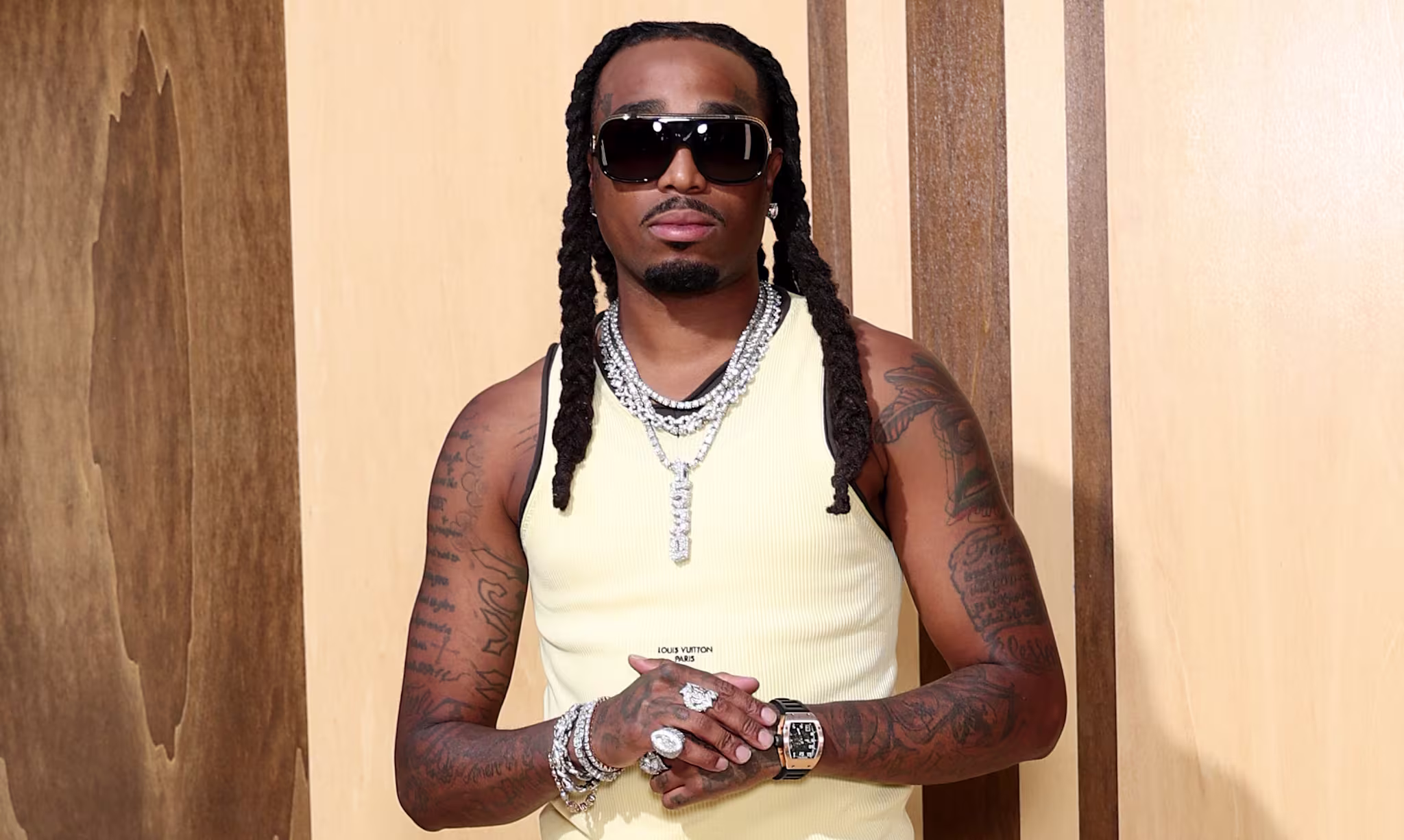 Quavo with long braids, wearing sunglasses, a yellow tank top, jeans, and orange sneakers, stands against a wooden background.
