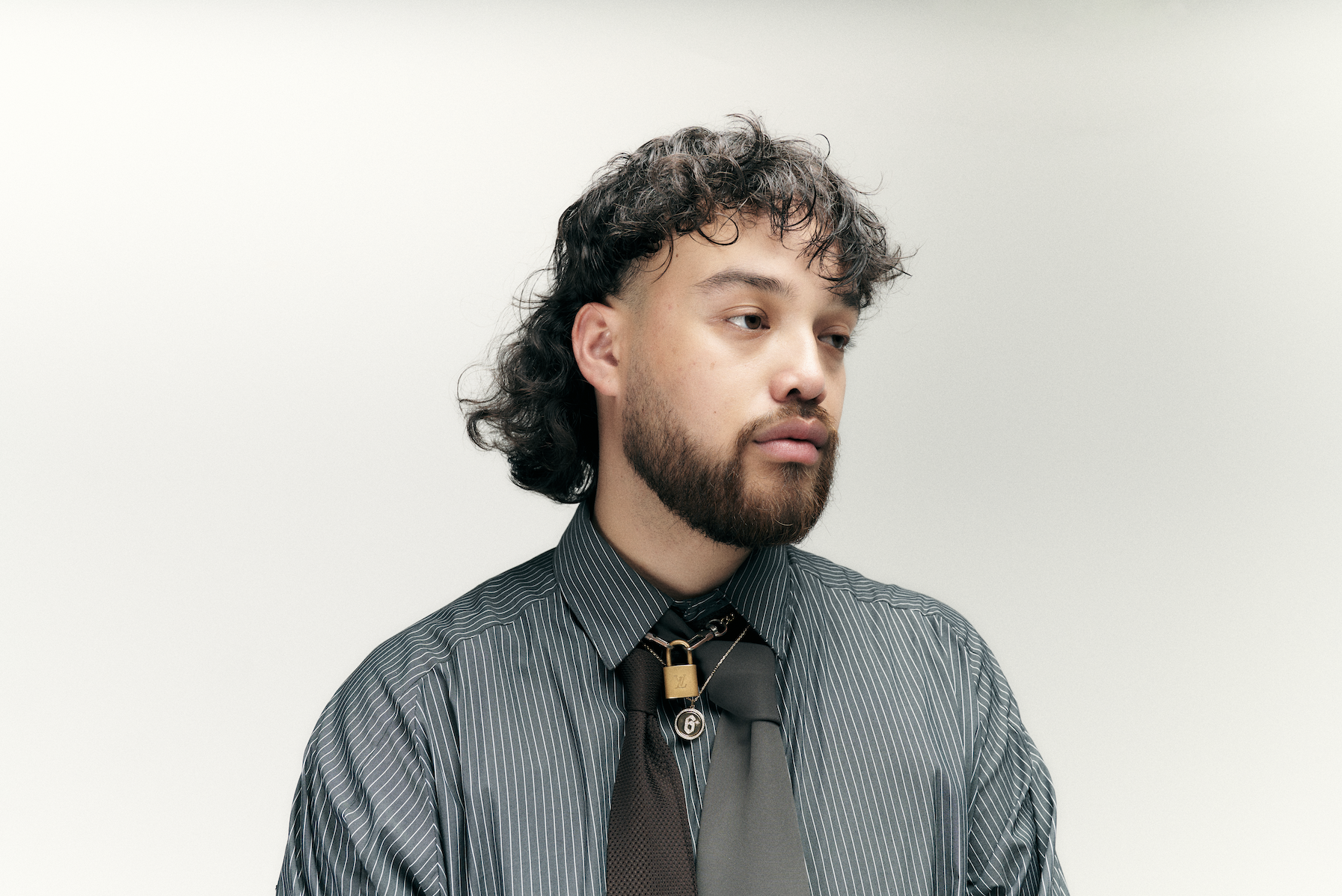 A man with curly hair and a beard wearing a striped shirt and tie, looking to the side against a plain background.
