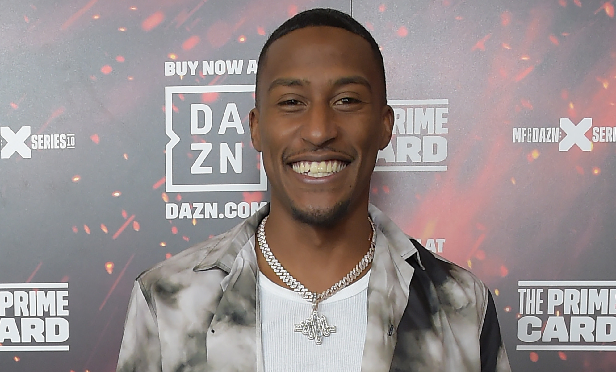 Yung Filly is smiling at an event, wearing a black and white shirt over a white tank top, with a necklace and watch.