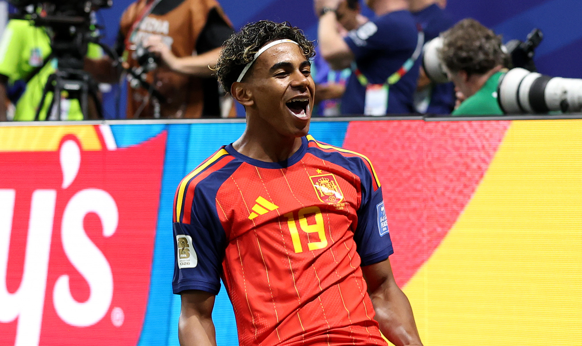 Lamine Yamal in a red and blue Spain jersey celebrates on the field, surrounded by photographers and an audience.