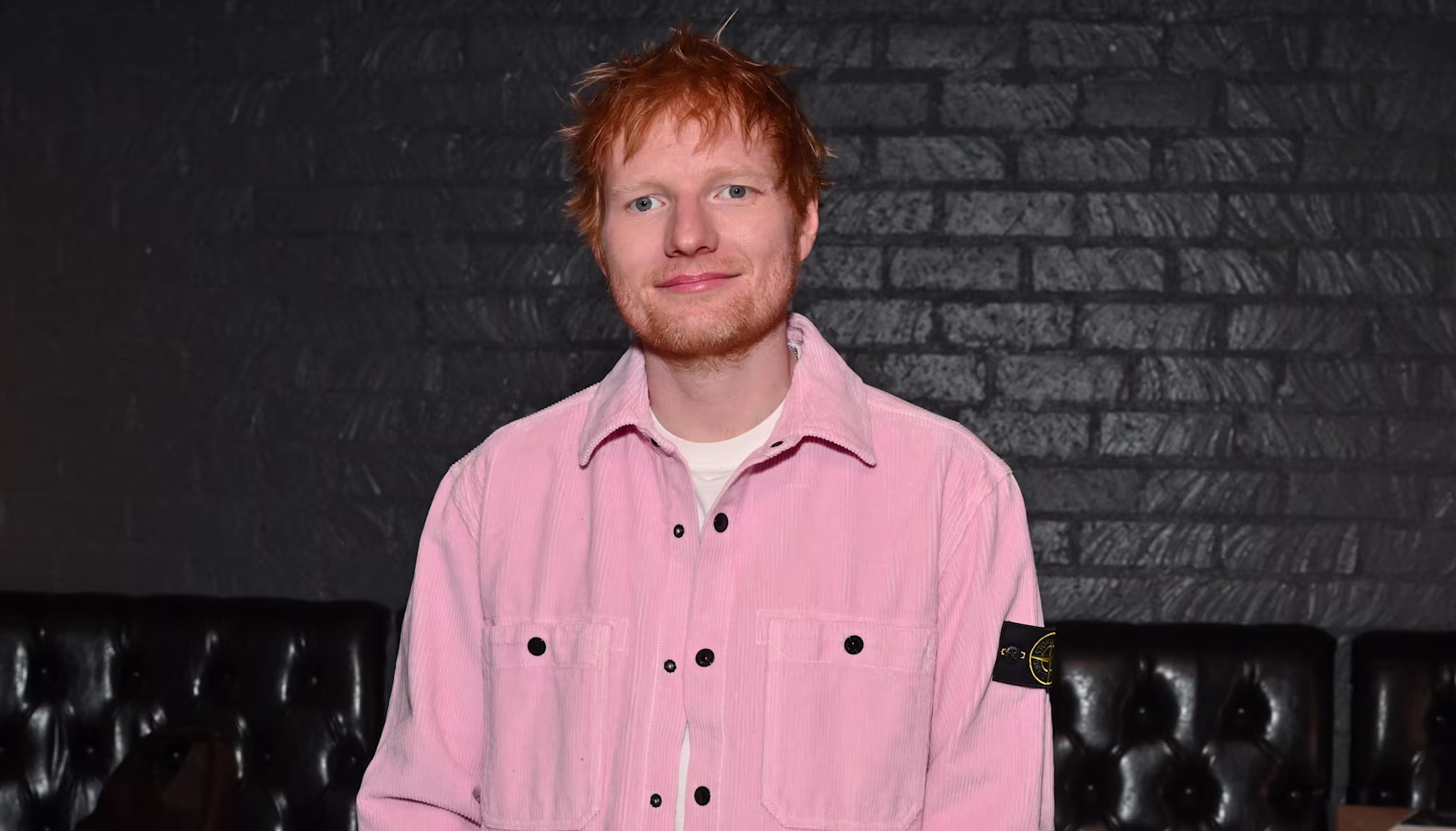 Ed Sheeran in a pink shirt, standing in a dimly lit room with black leather seating and equipment in the background.