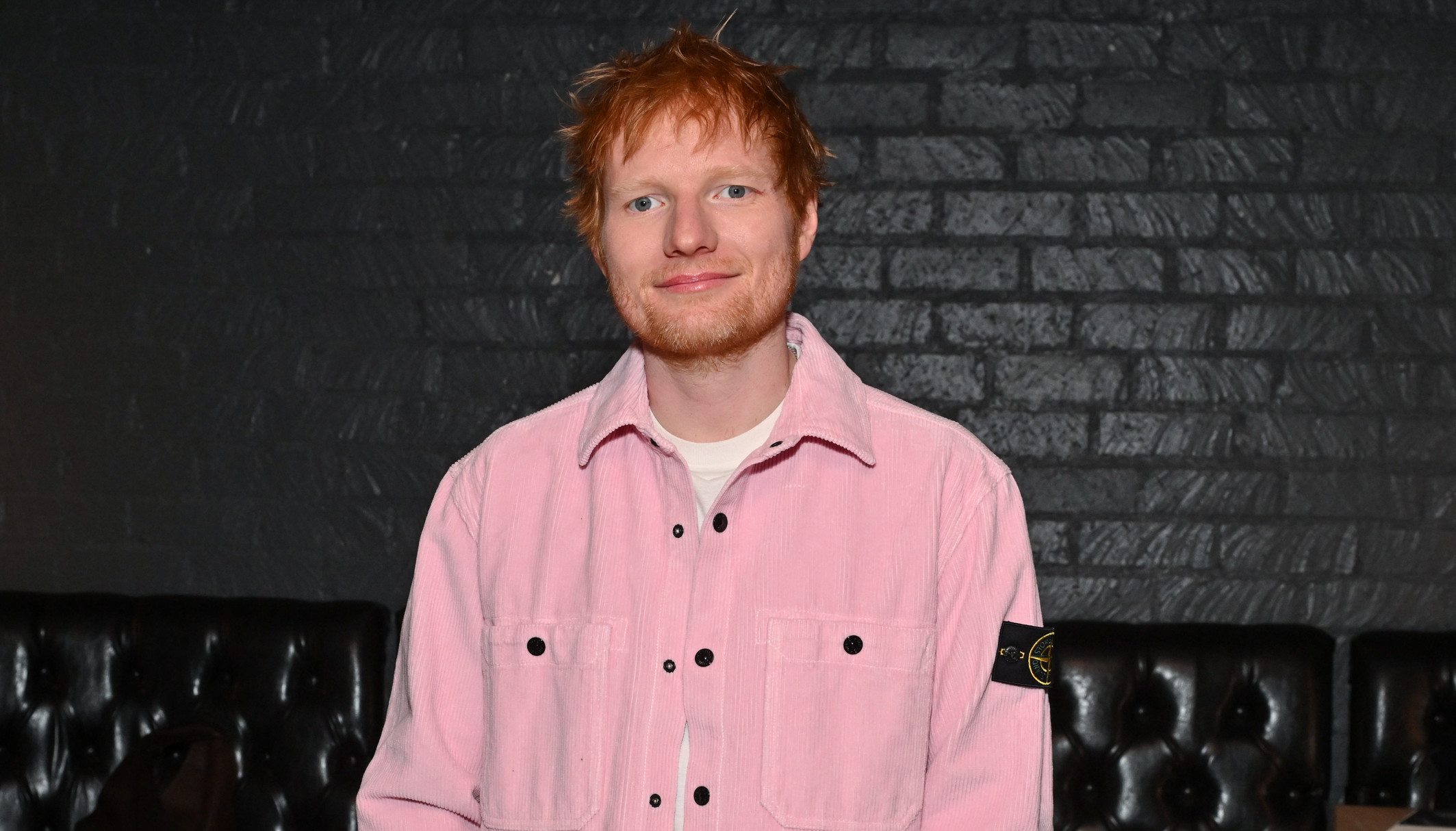 Ed Sheeran in a pink shirt, standing in a dimly lit room with black leather seating and equipment in the background.