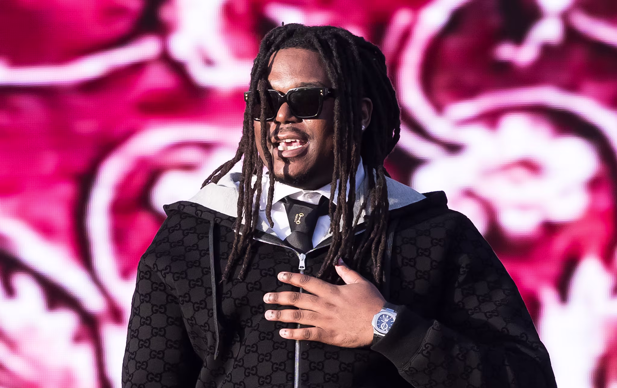 LUCKI with dreadlocks and sunglasses performs on stage, wearing a black tracksuit, against a red and white patterned background.