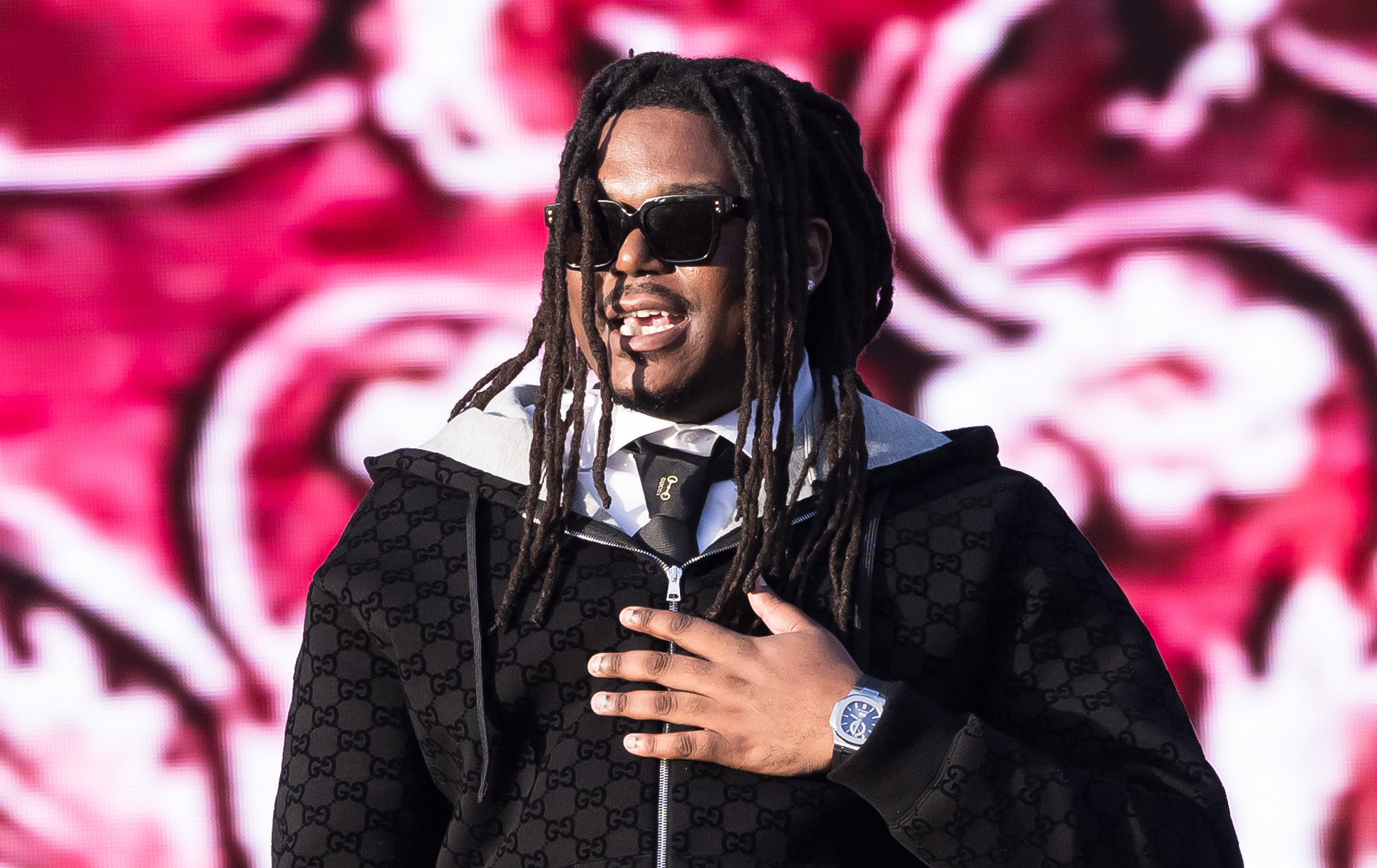 LUCKI with dreadlocks and sunglasses on stage, wearing a black tracksuit, holding a microphone against a vibrant red and white backdrop.