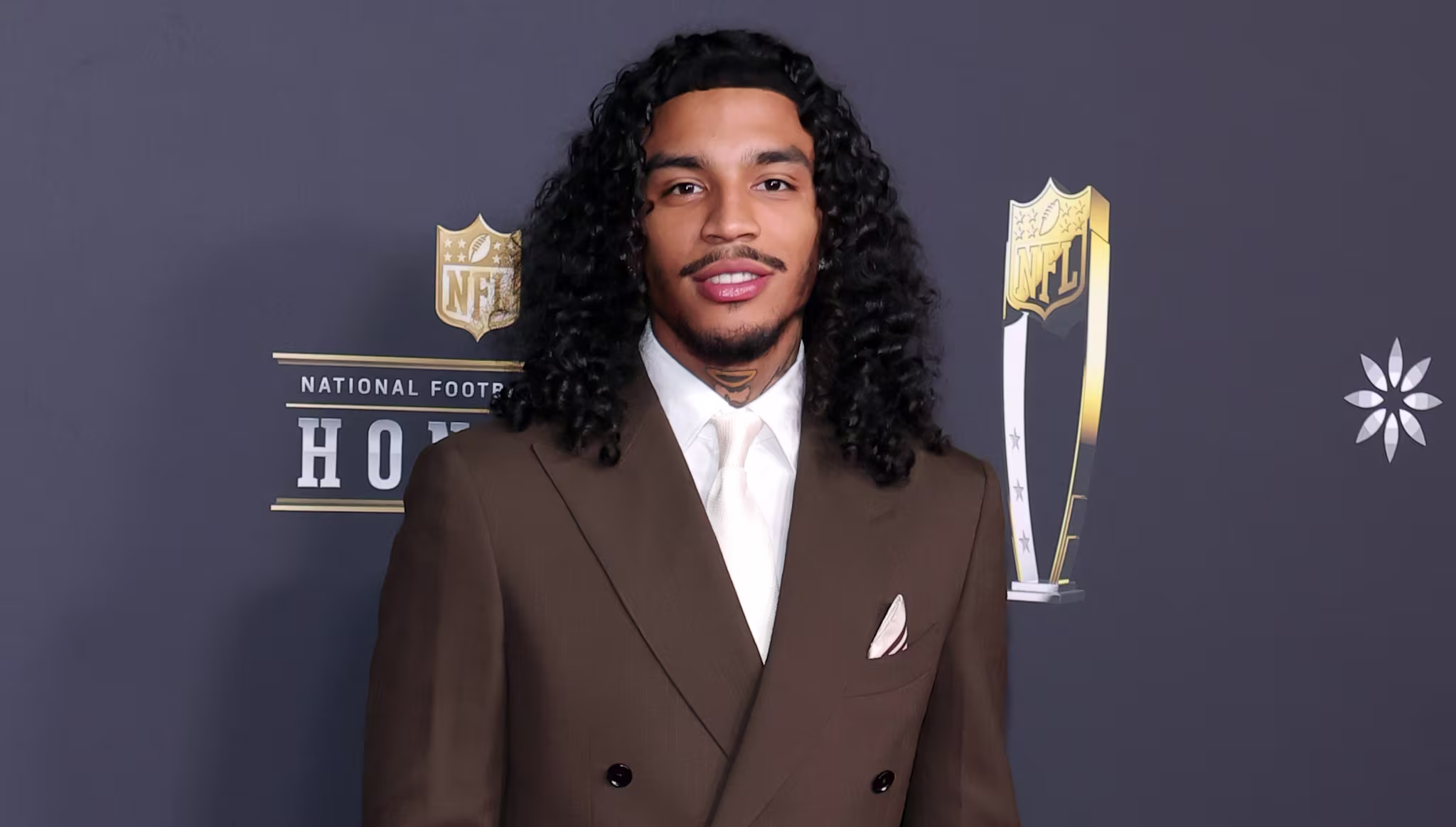Jay Cinco in a brown suit with long curly hair stands on the red carpet at the NFL Honors event.