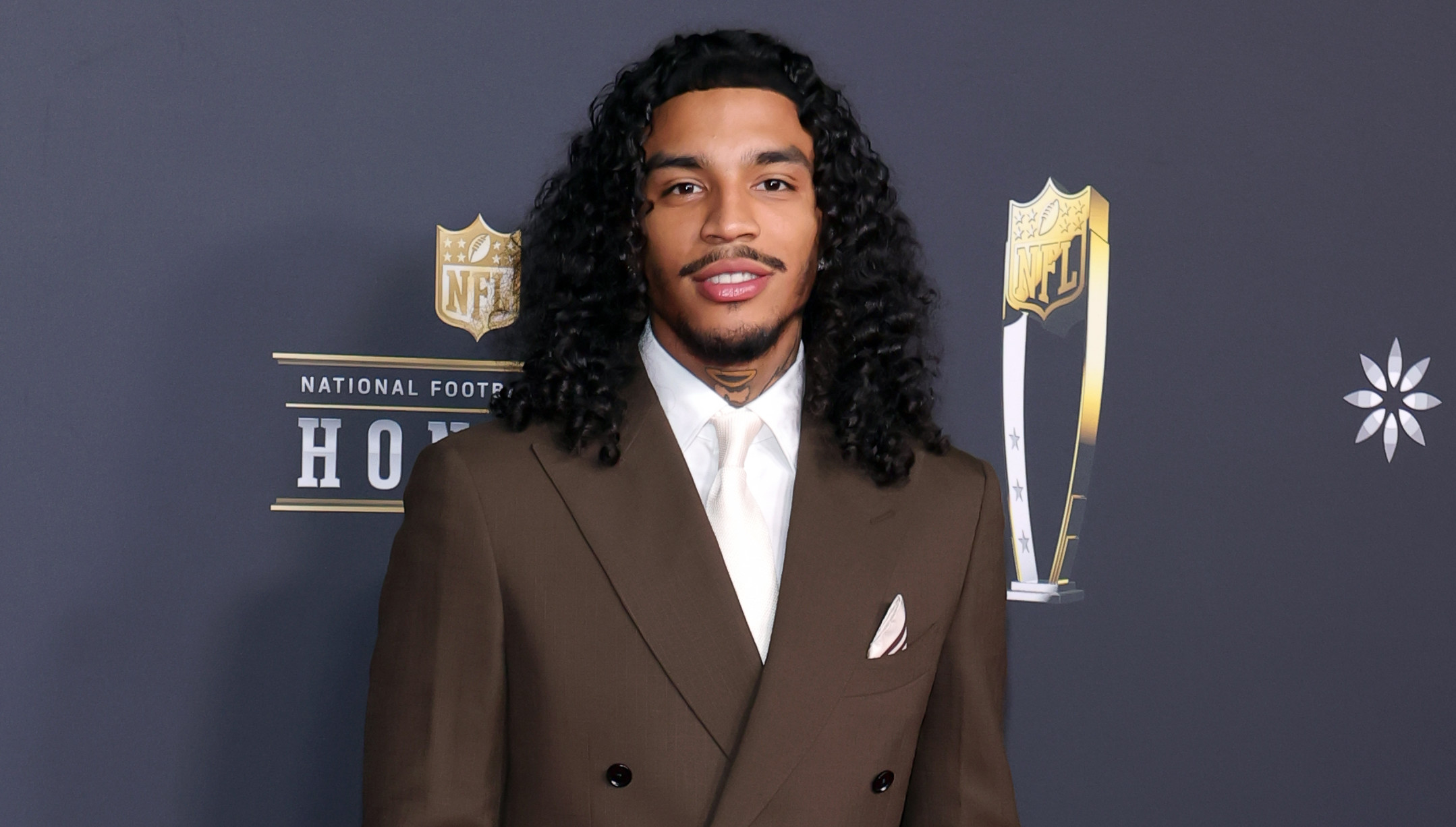 Jay Cinco in a brown suit with long curly hair stands on the red carpet at the NFL Honors event.