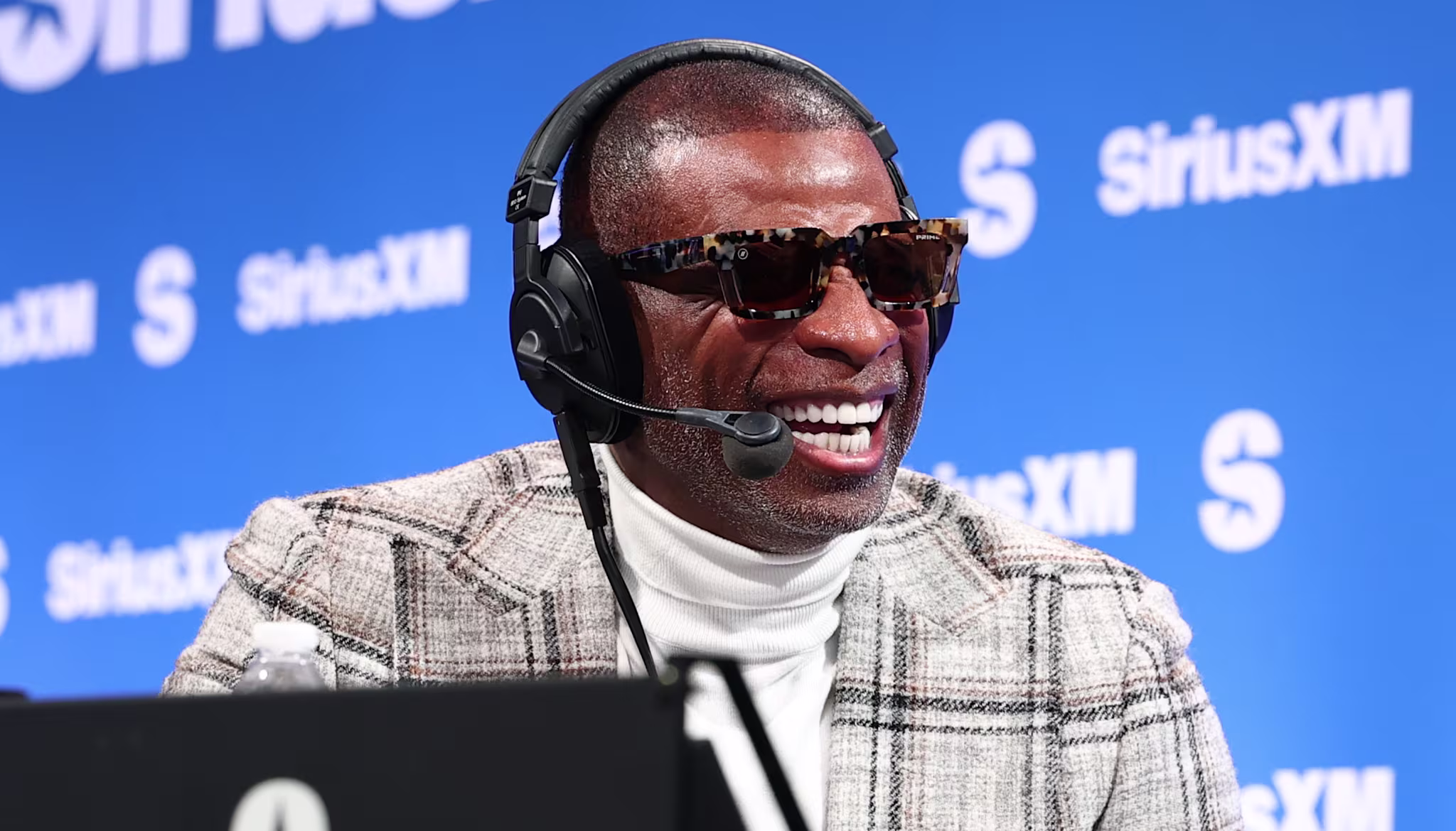 Deion Sanders wearing sunglasses and a headset, smiling while broadcasting at a SiriusXM event.