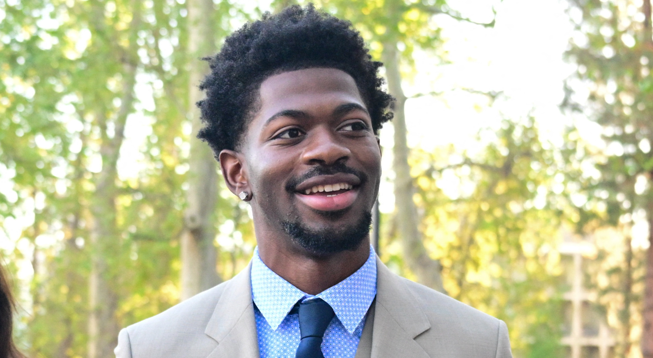 Lil Nas X in a beige suit and blue tie, smiling outdoors with a woman in a blue blazer and glasses. Trees in the background.