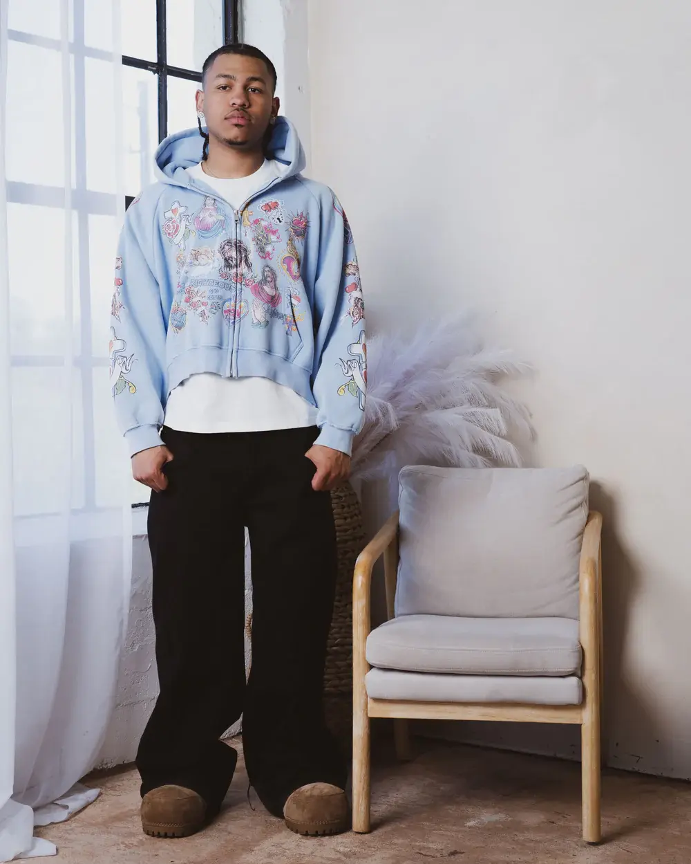 A person in a blue hoodie with colorful designs, black pants, and brown shoes stands next to a light-colored chair indoors.